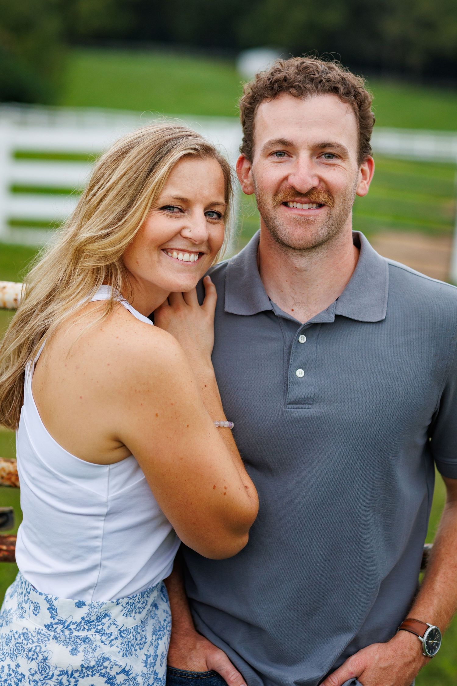 Smiling couple posing together near a white fence on a farm, woman leaning affectionately against mans shoulder.
