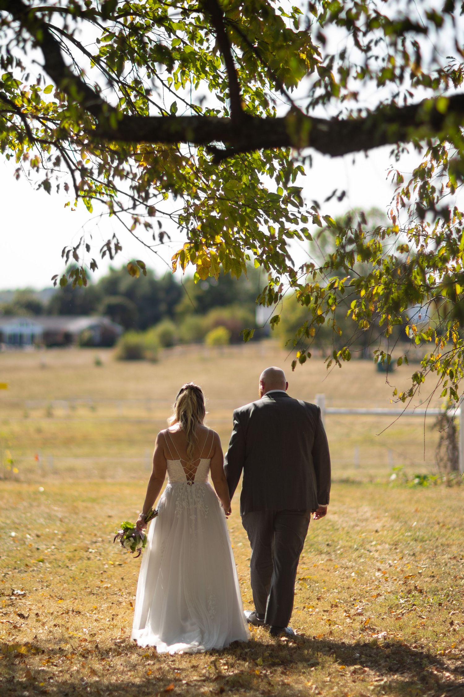 Bride and groom walking together through a sun-drenched autumn field beneath golden tree branches.