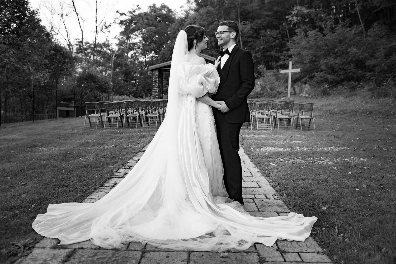 Bride and groom pose on brick path with long veil trailing behind in black and white portrait.