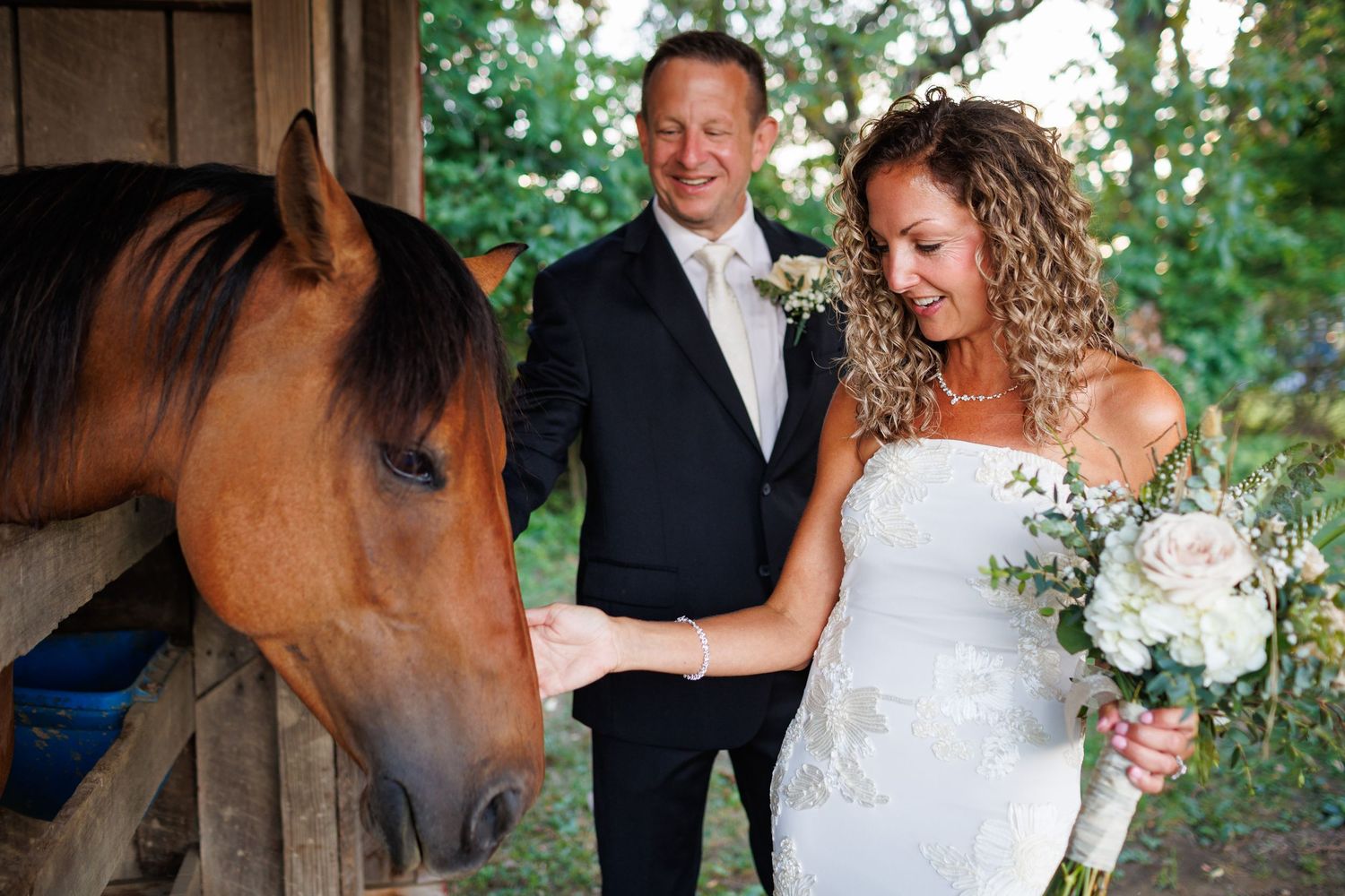 Bride and groom smiling while petting a brown horse at a rustic barn wedding venue.