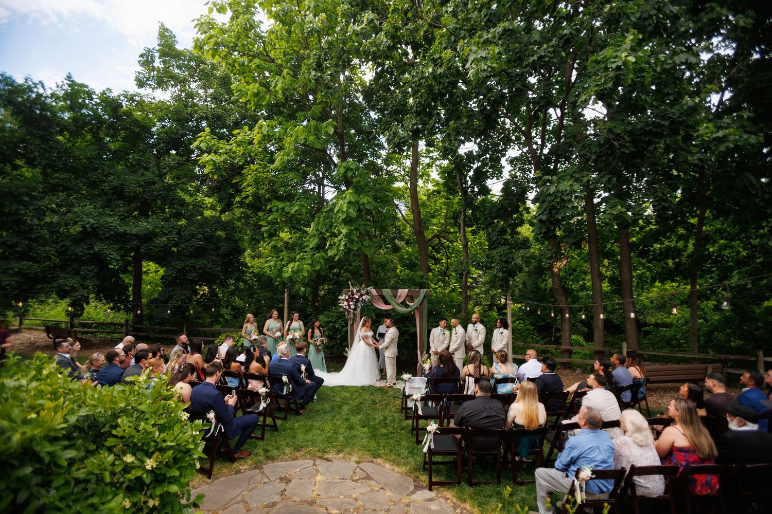 Outdoor wedding ceremony in lush garden setting with guests seated and couple at floral arch.