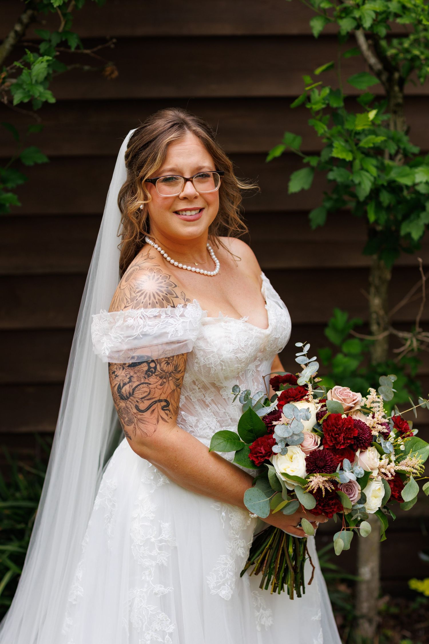 Smiling bride in off-shoulder lace gown with veil, pearl necklace, and red and blush bridal bouquet outdoors.