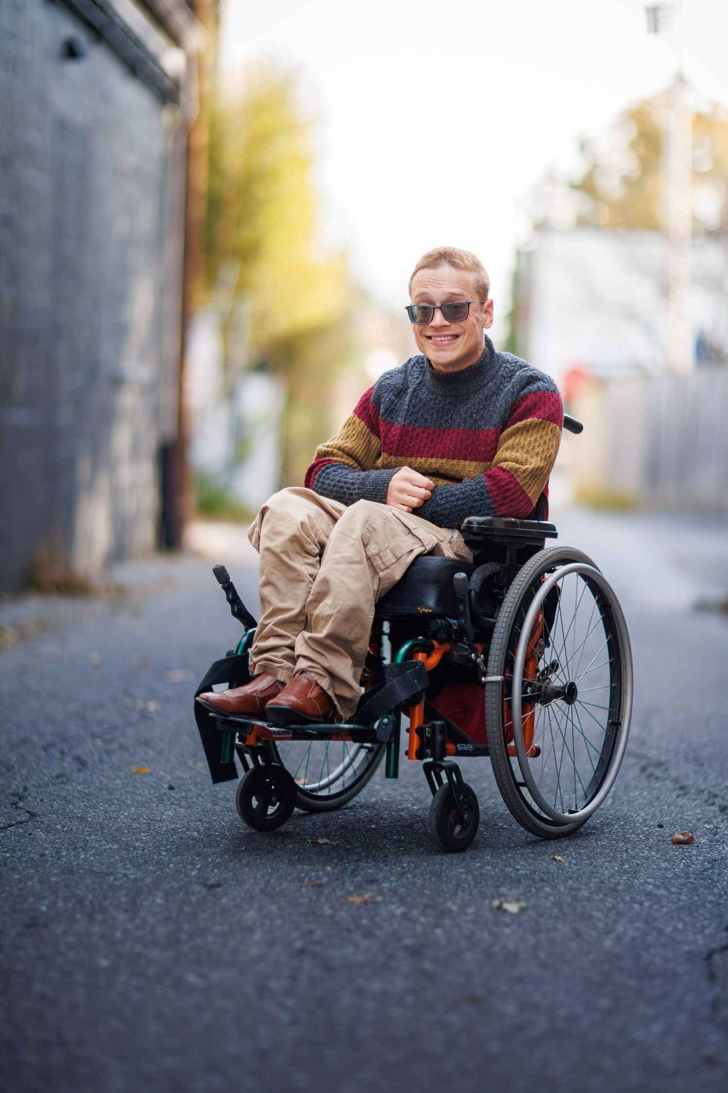 Person in a colorful sweater and sunglasses smiles while sitting in a wheelchair on an autumn street.