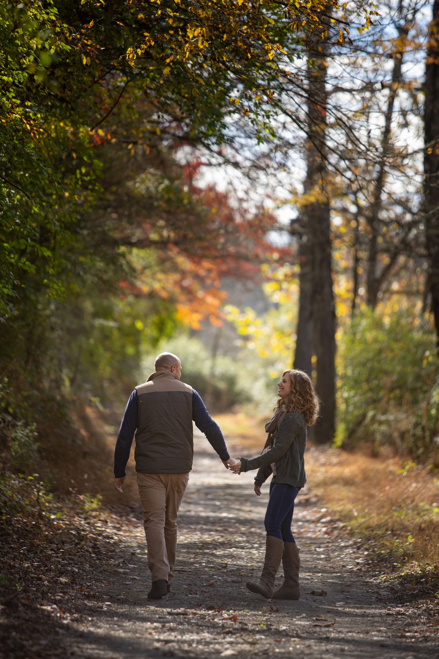 Two people walk hand in hand down a sunlit autumn forest path surrounded by golden leaves and trees.