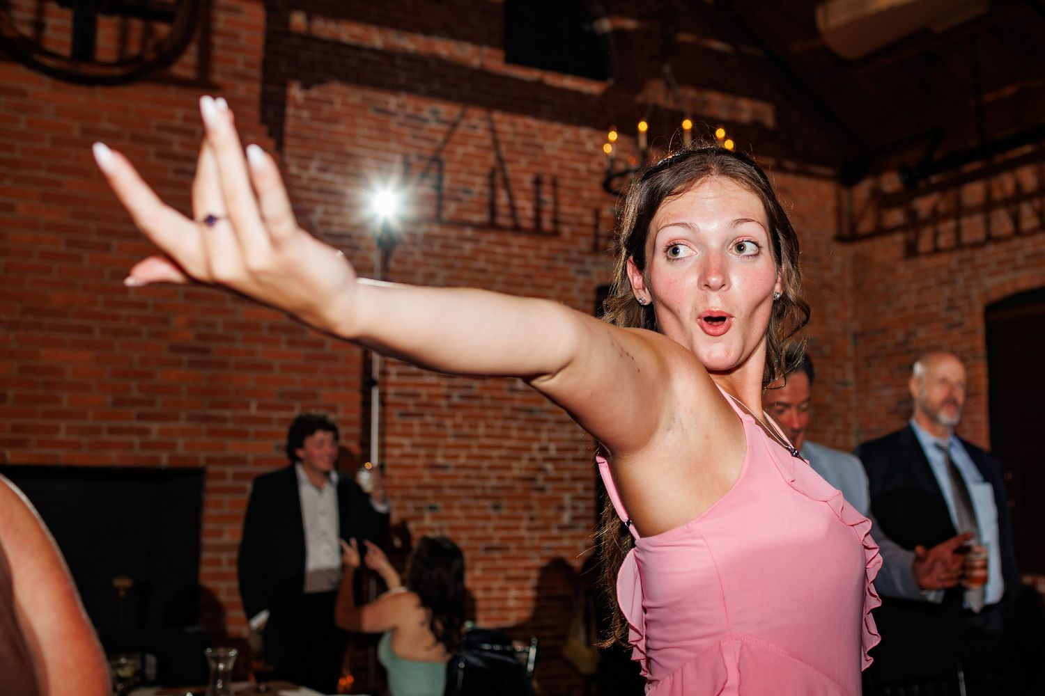 Excited woman in pink dress dancing energetically at a wedding reception in a brick venue.