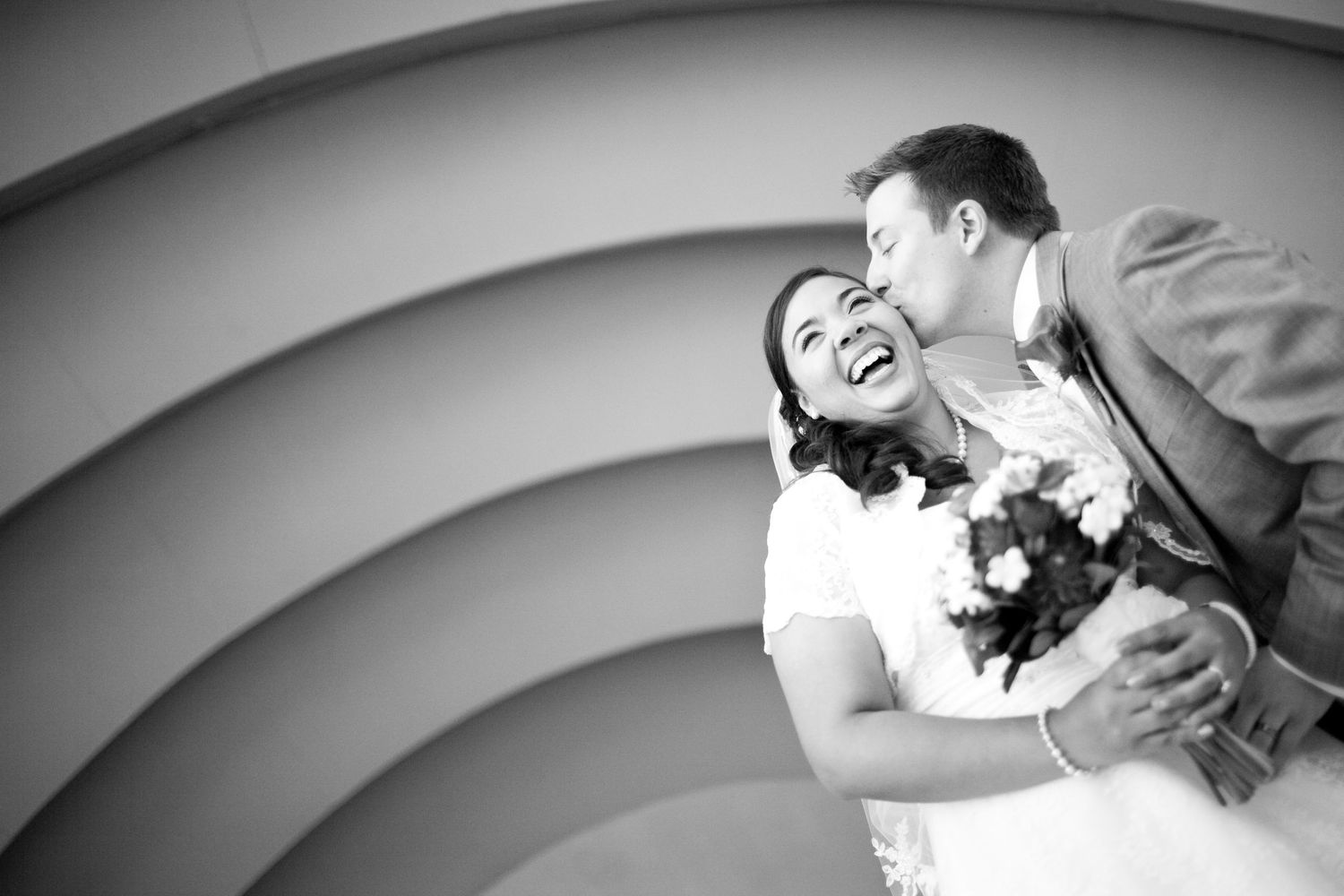 Black and white romantic photo of a couple laughing together holding flowers against a curved background.