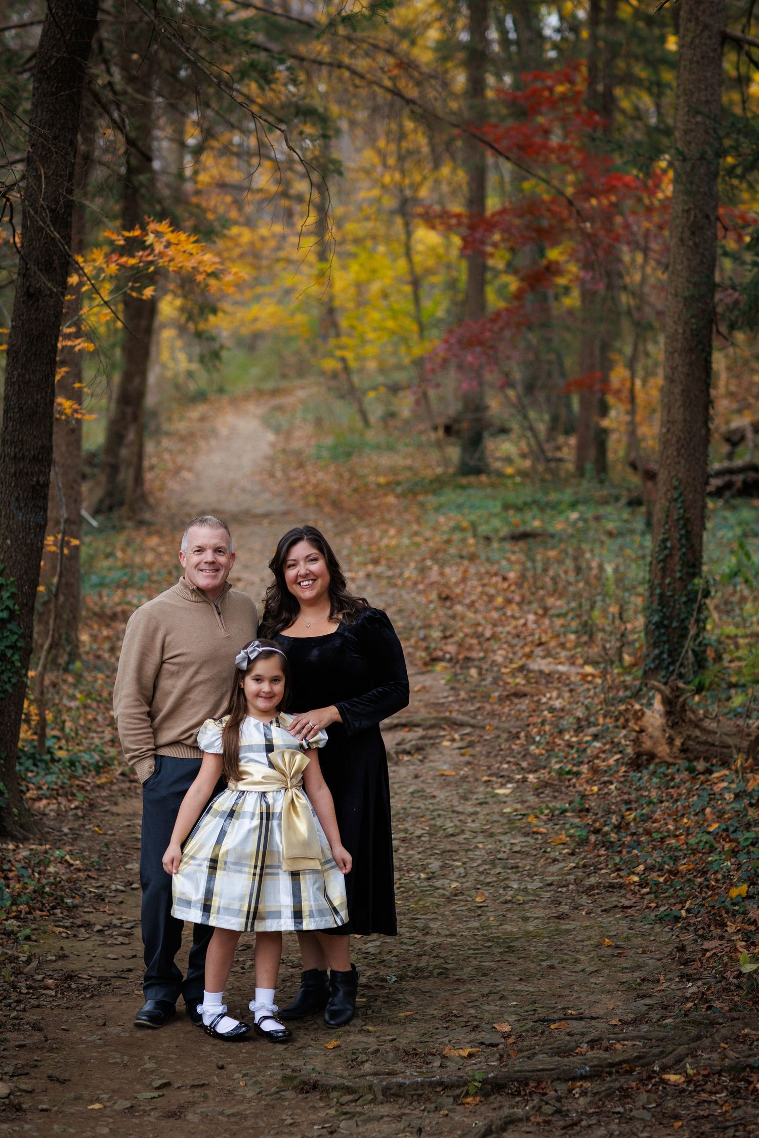 A family poses together on a leaf-covered forest path during autumn with colorful fall foliage in the background.