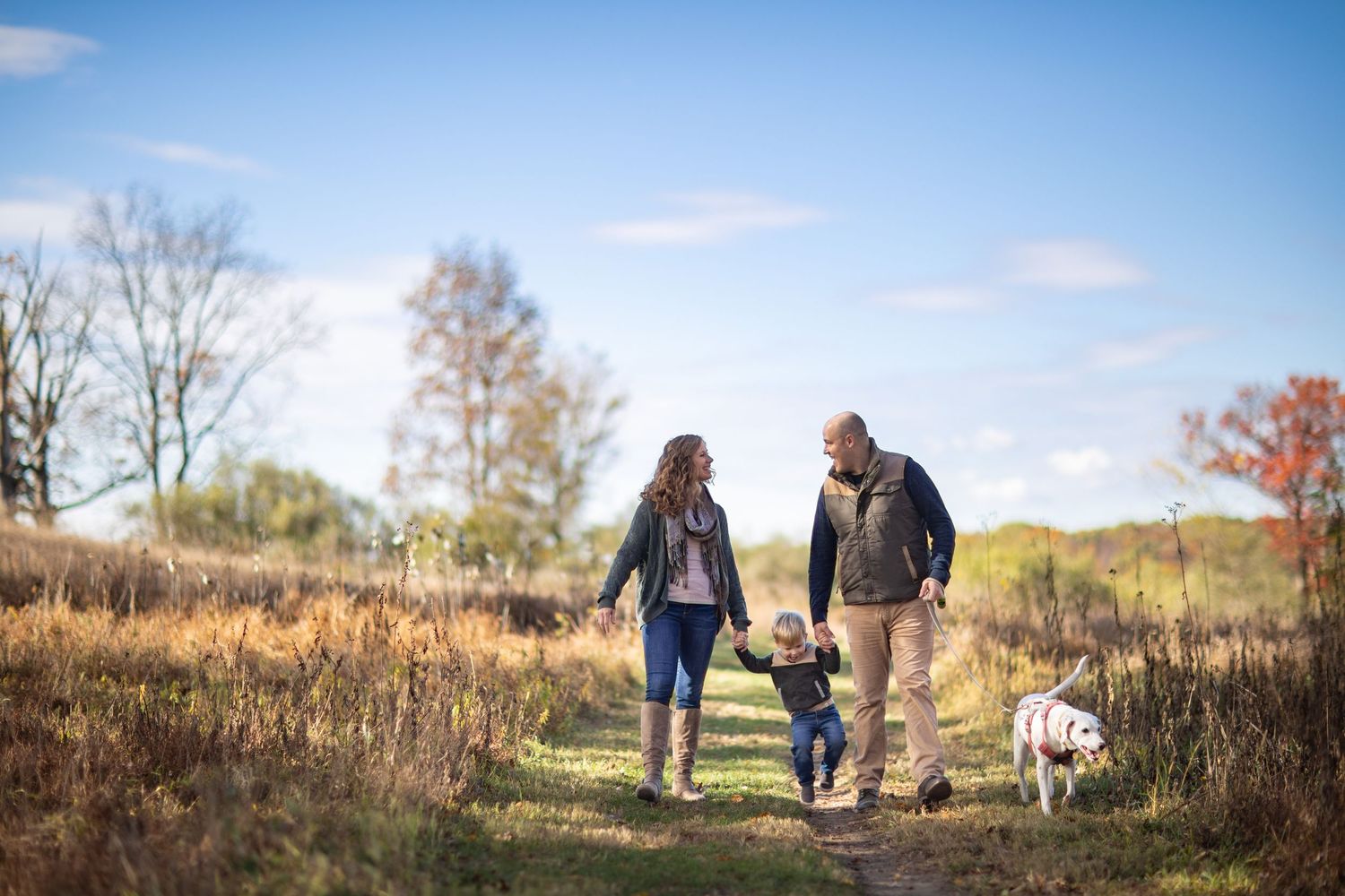 A couple walks with their dog through a scenic autumn field during a golden sunset.