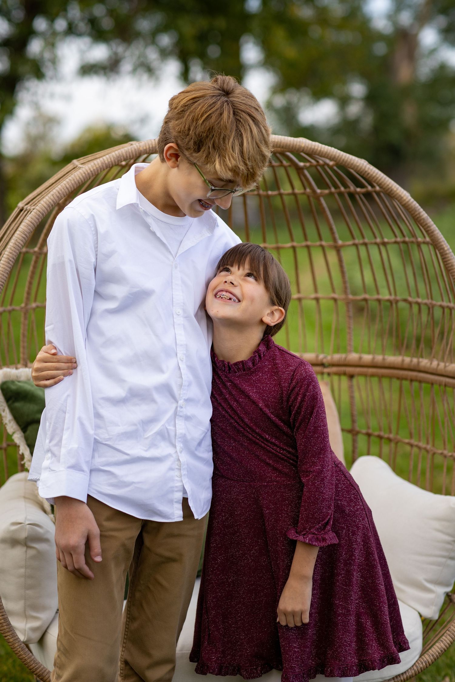 Two children in white and burgundy outfits share a playful moment outdoors near a wooden fence.