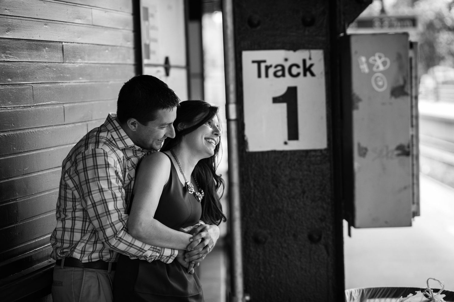 Romantic black and white photo of couple embracing on train platform near Track 1 sign.