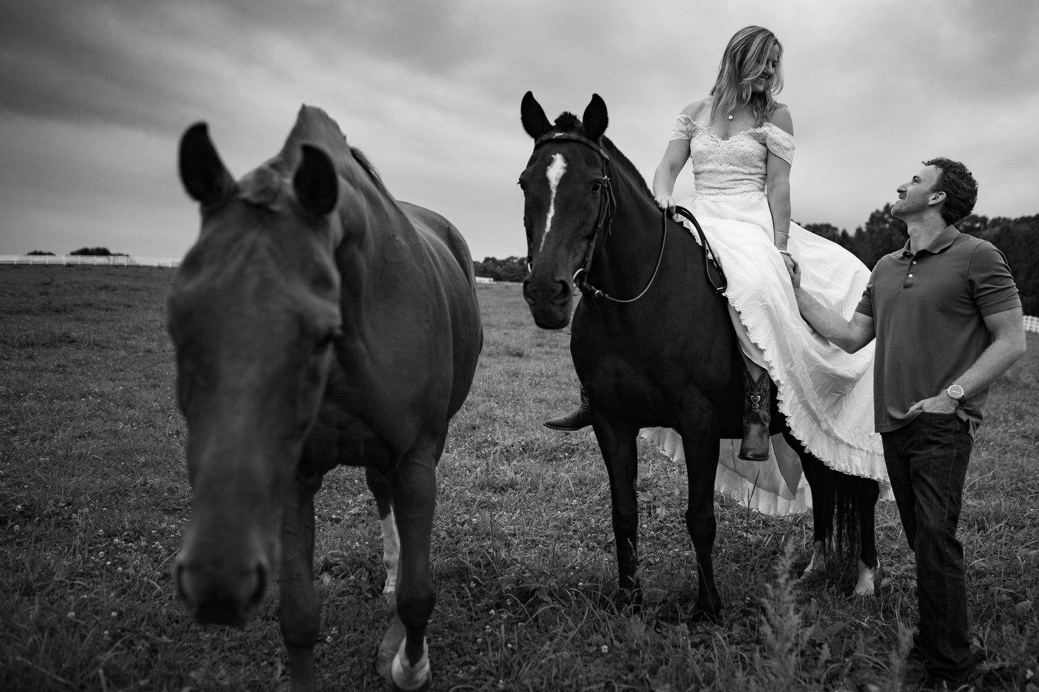 Black and white photo of a bride on horseback with groom standing beside her in a field.