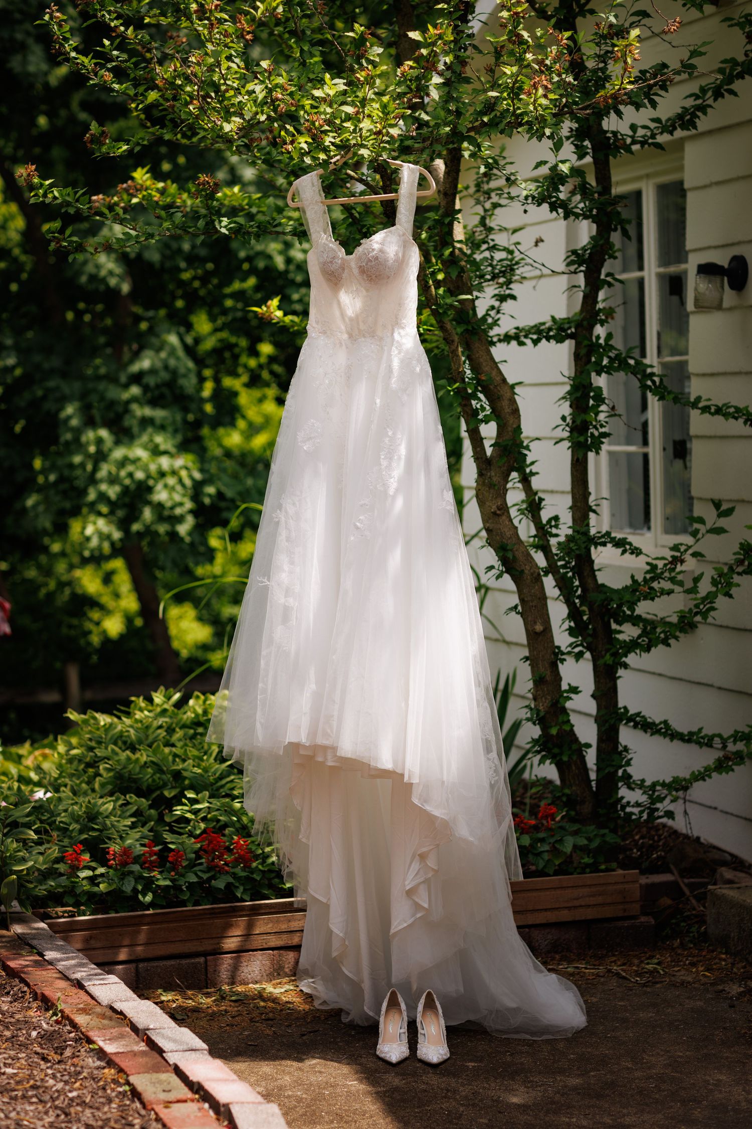 Elegant white lace wedding gown hanging outdoors on a tree with matching bridal heels below.