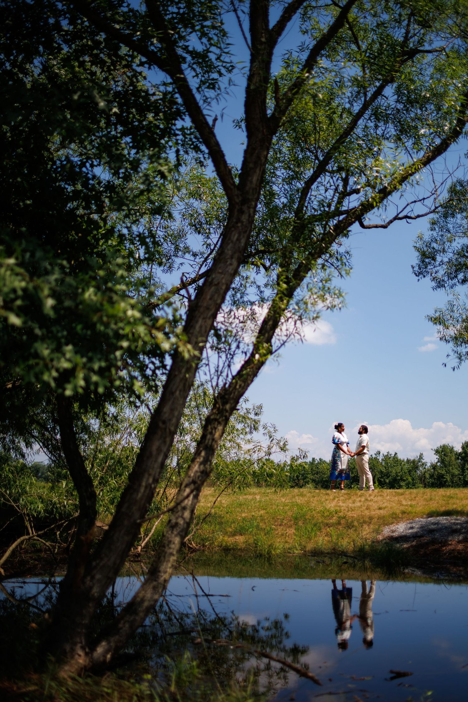 Couple holding hands reflected in pond water, framed by lush green trees under blue sky in scenic outdoor setting.