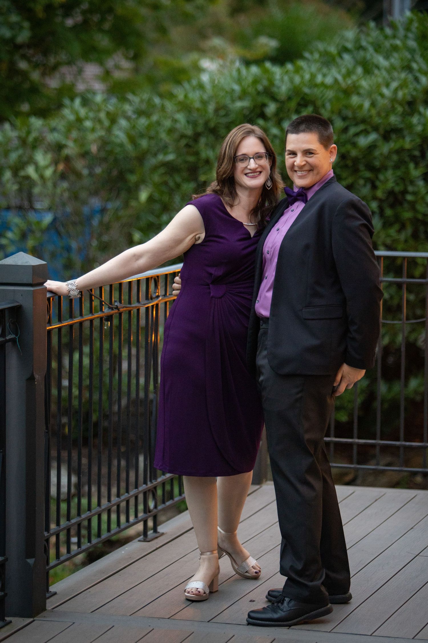 A couple in formal attire poses playfully on a wooden deck surrounded by greenery.