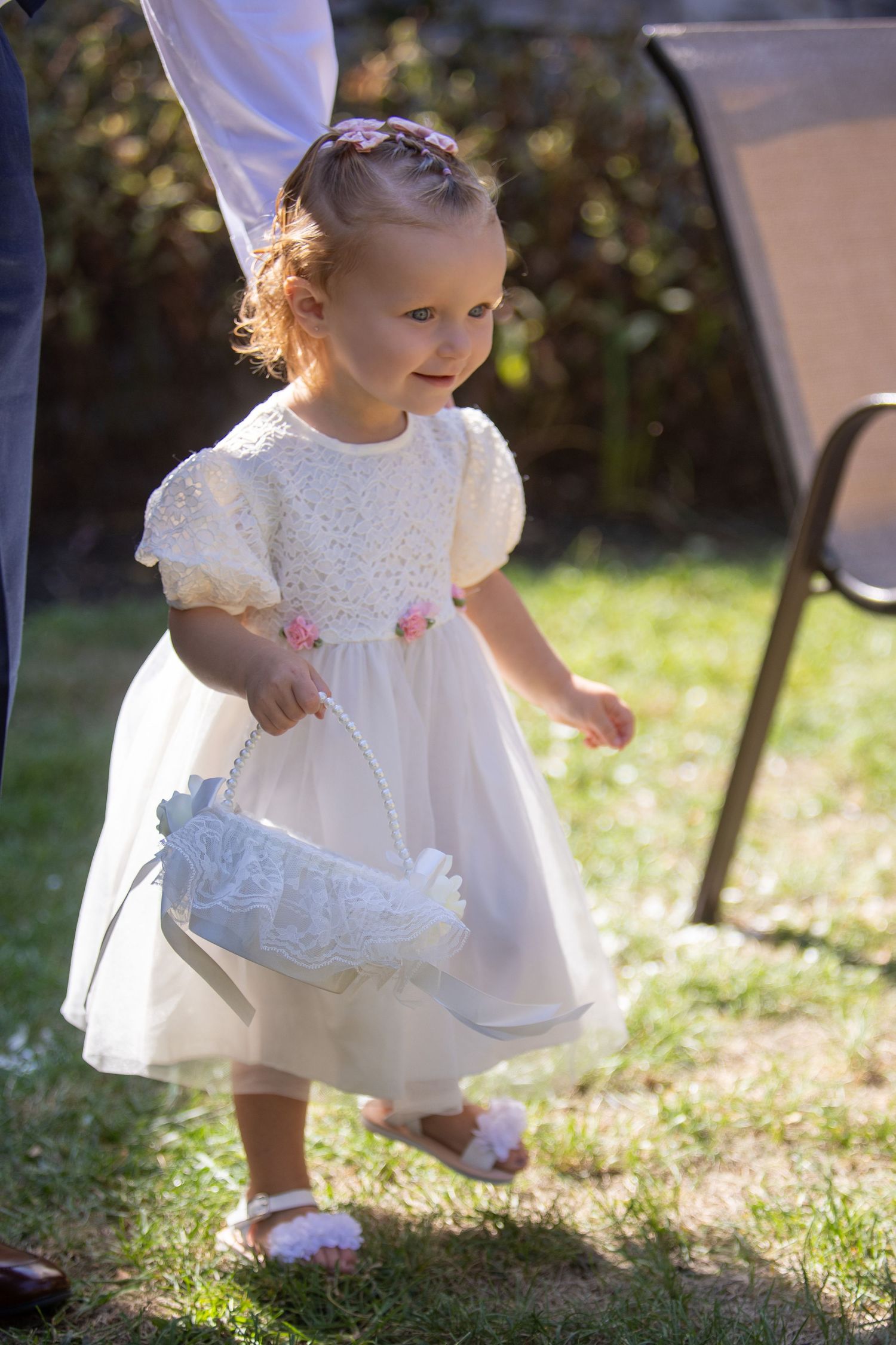 A toddler in a white lace dress plays on the grass during a sunny day outdoors.