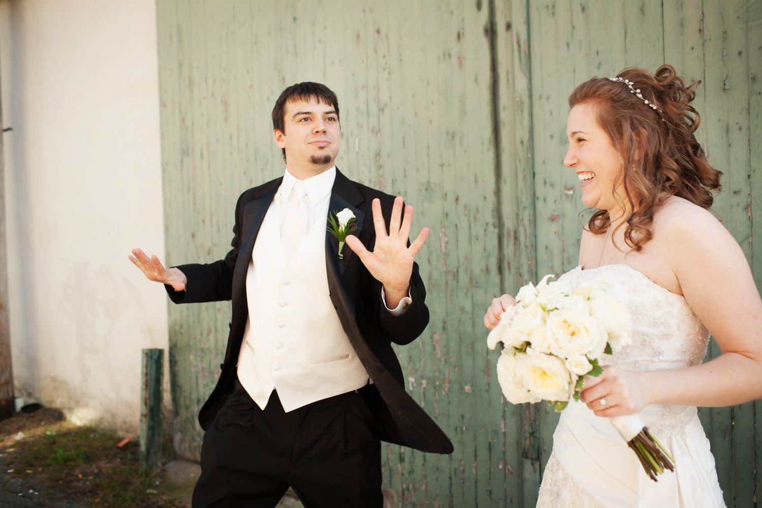 A groom in a tuxedo is dancing while the bride smiles and laughs. They are in an alley