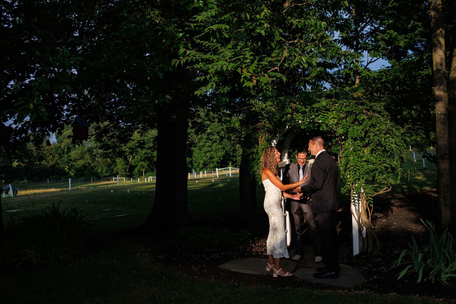 Couple exchanges vows outdoors under lush green trees at golden hour with an officiant at an intimate garden wedding ceremony.