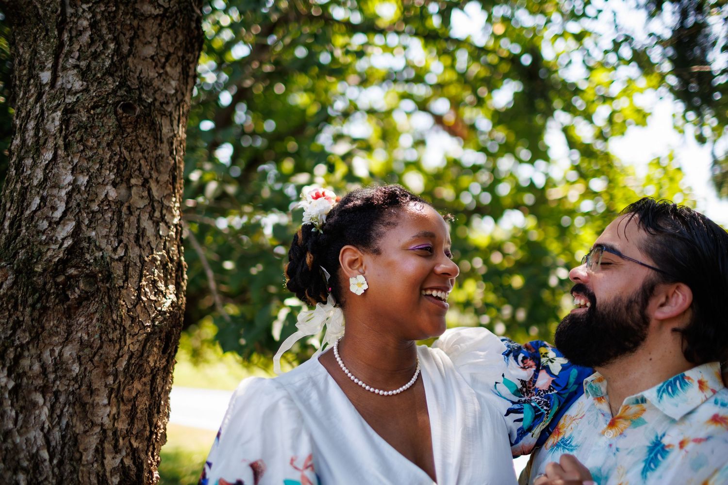 Happy interracial couple laughing together outdoors near a tree, bride in white dress with pearl necklace and floral hair accessory.