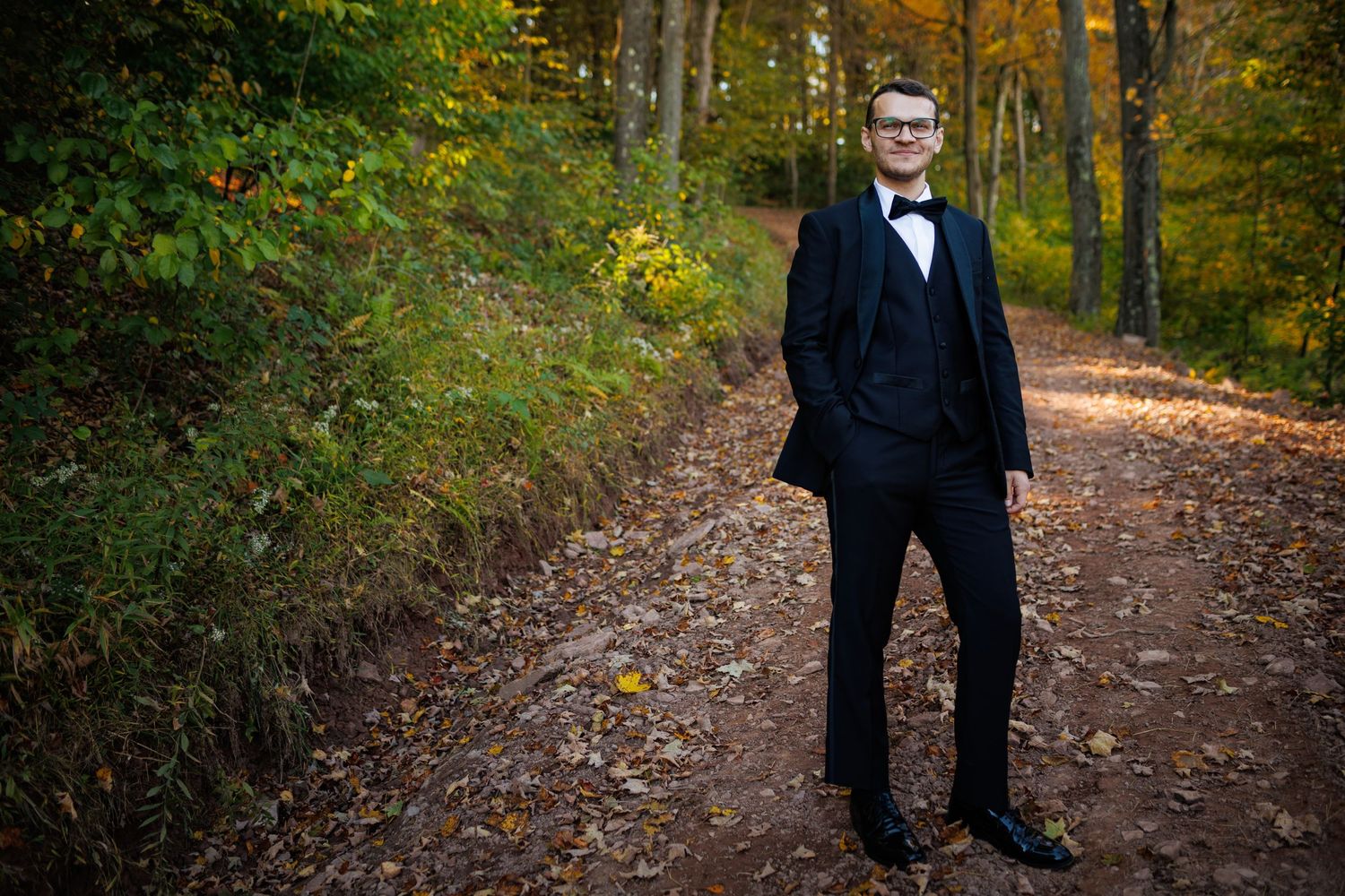 Groom in black tuxedo poses confidently on autumn forest path with colorful foliage.