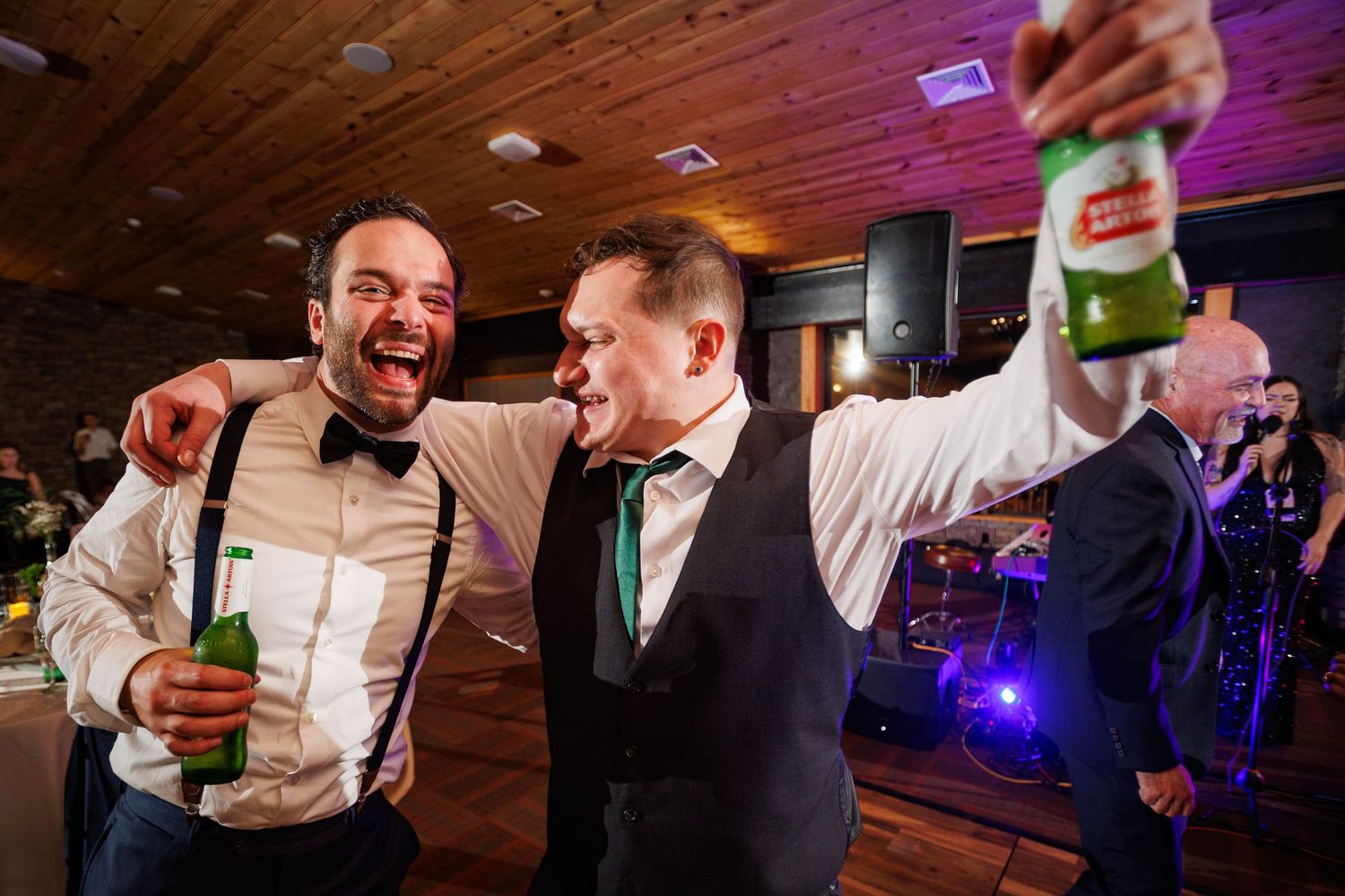 Two groomsmen celebrate joyfully on wedding dance floor holding green beer bottles.