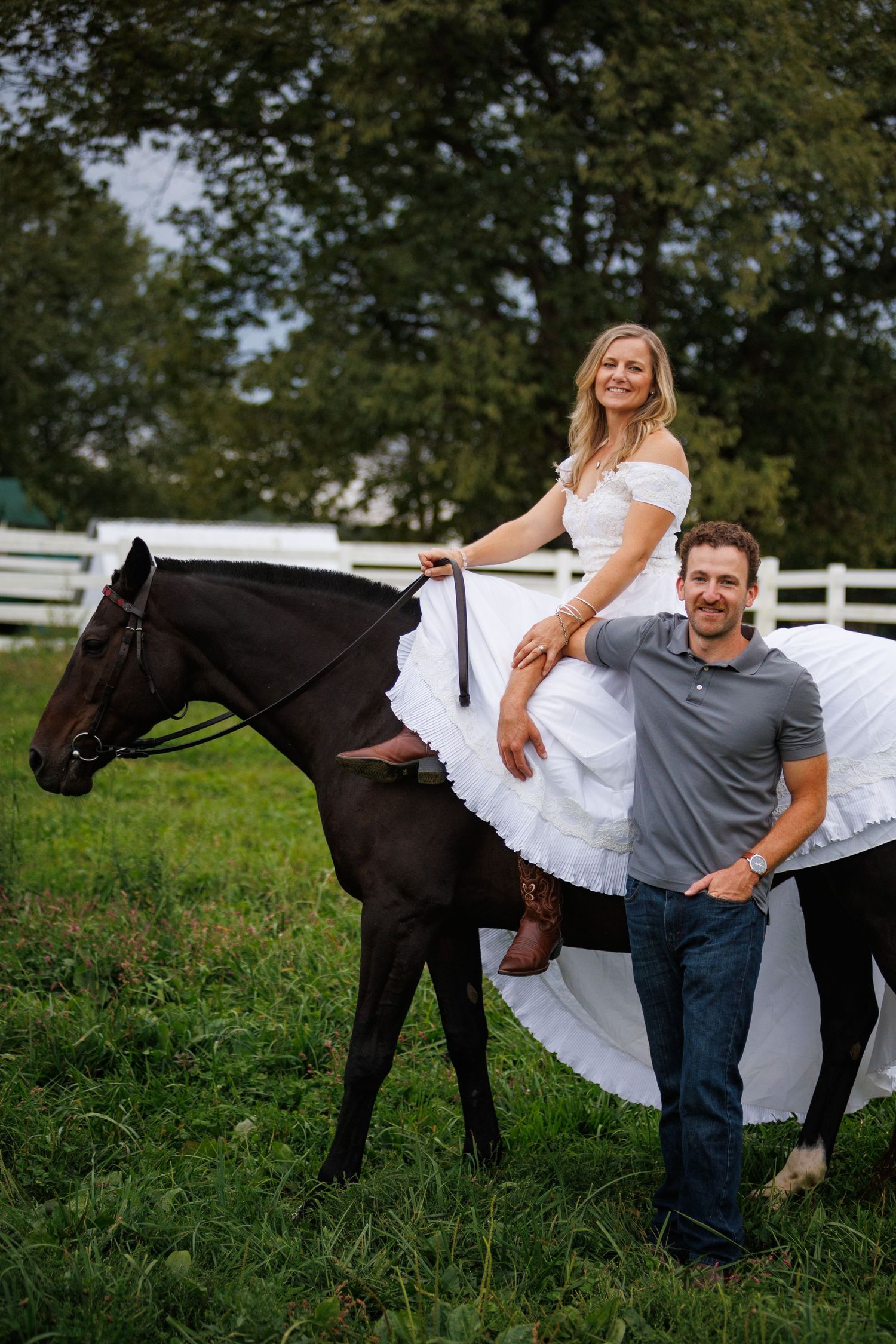 A smiling blonde woman in a white dress sits on a dark horse beside a man in a gray polo and jeans on a green farm.