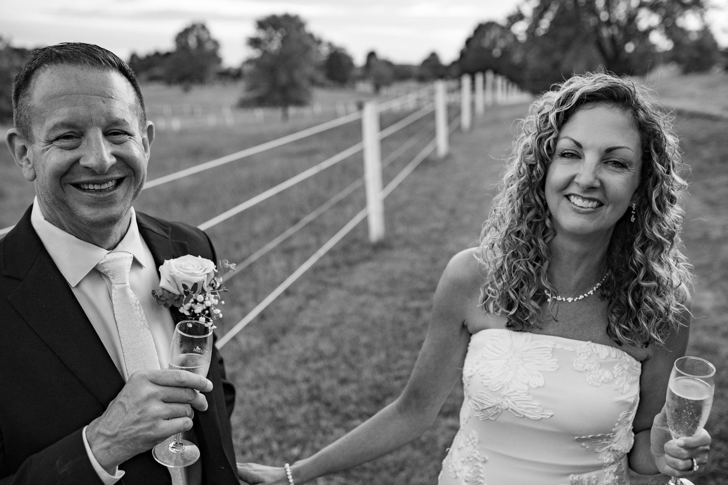 Smiling bride and groom holding champagne and bouquet beside white fence at outdoor wedding celebration.