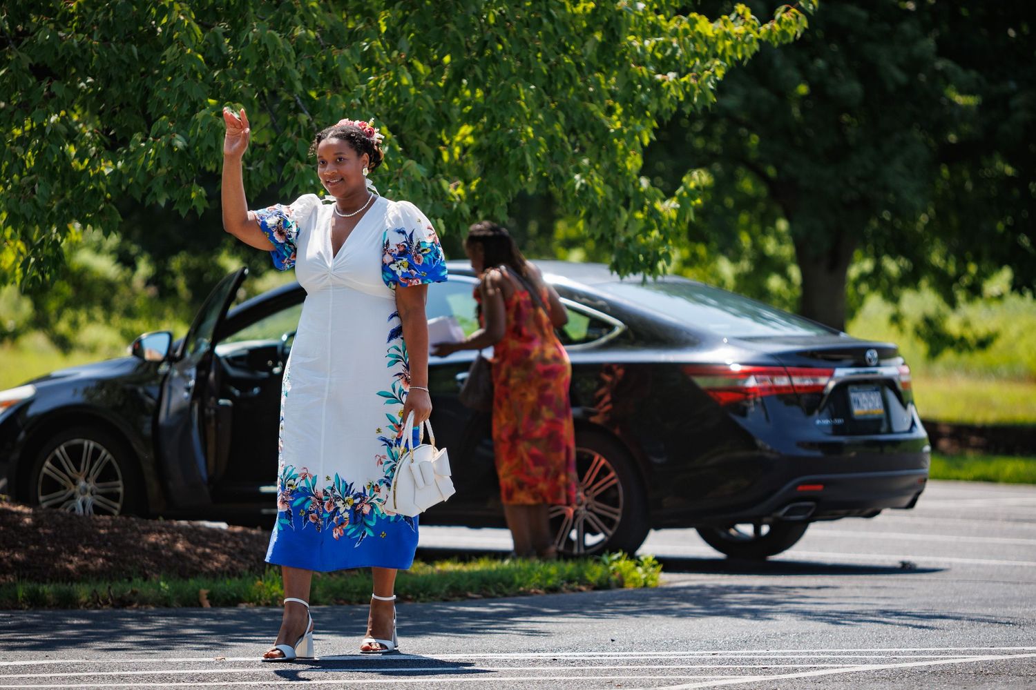 Woman in white floral dress waving in parking lot, another woman in red dress near black sedan on sunny day.