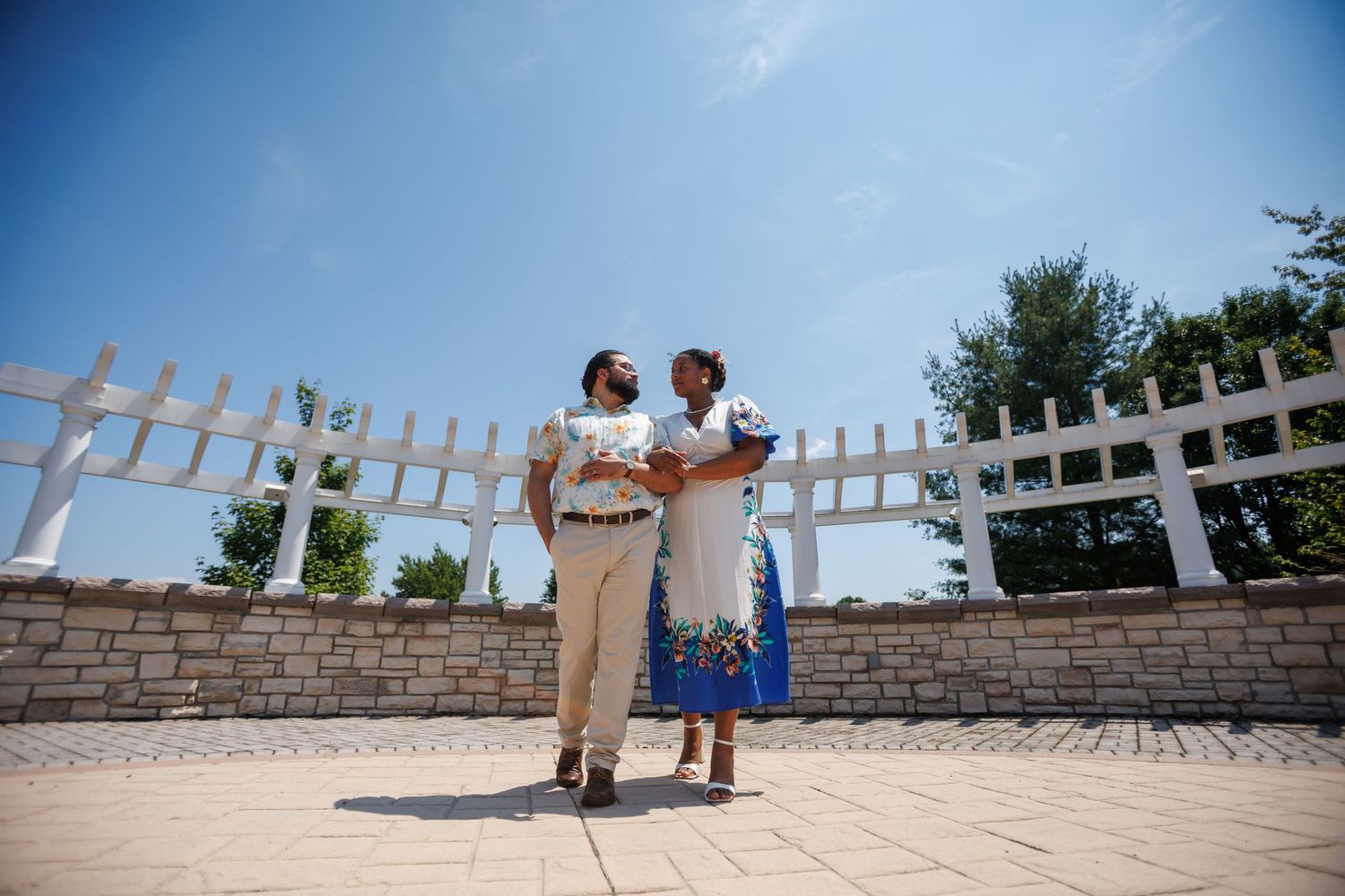 Couple in formal attire standing together in an outdoor courtyard with white lattice fence under a clear blue sky.