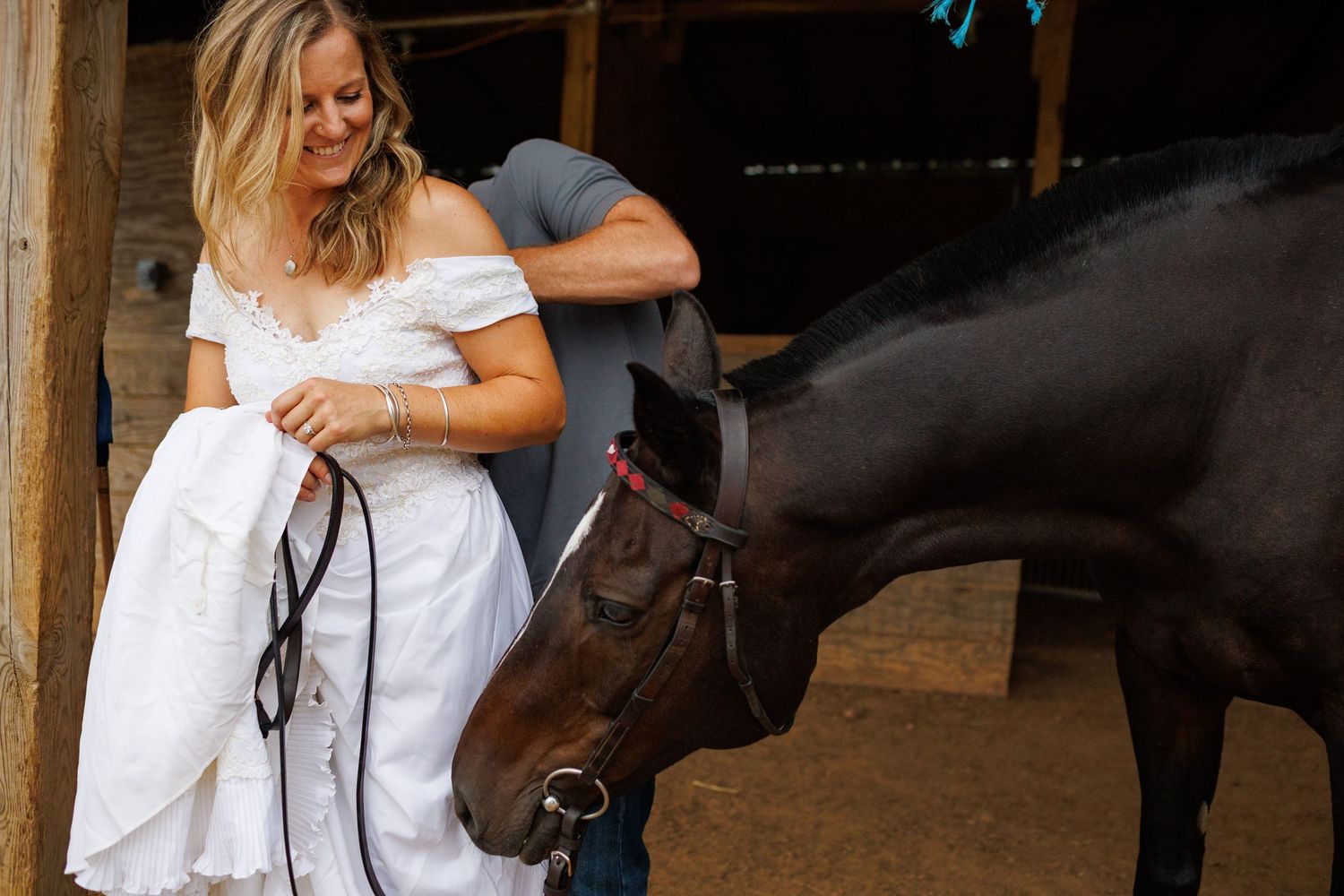 Bride in white dress smiling at a dark horse in a rustic barn setting.