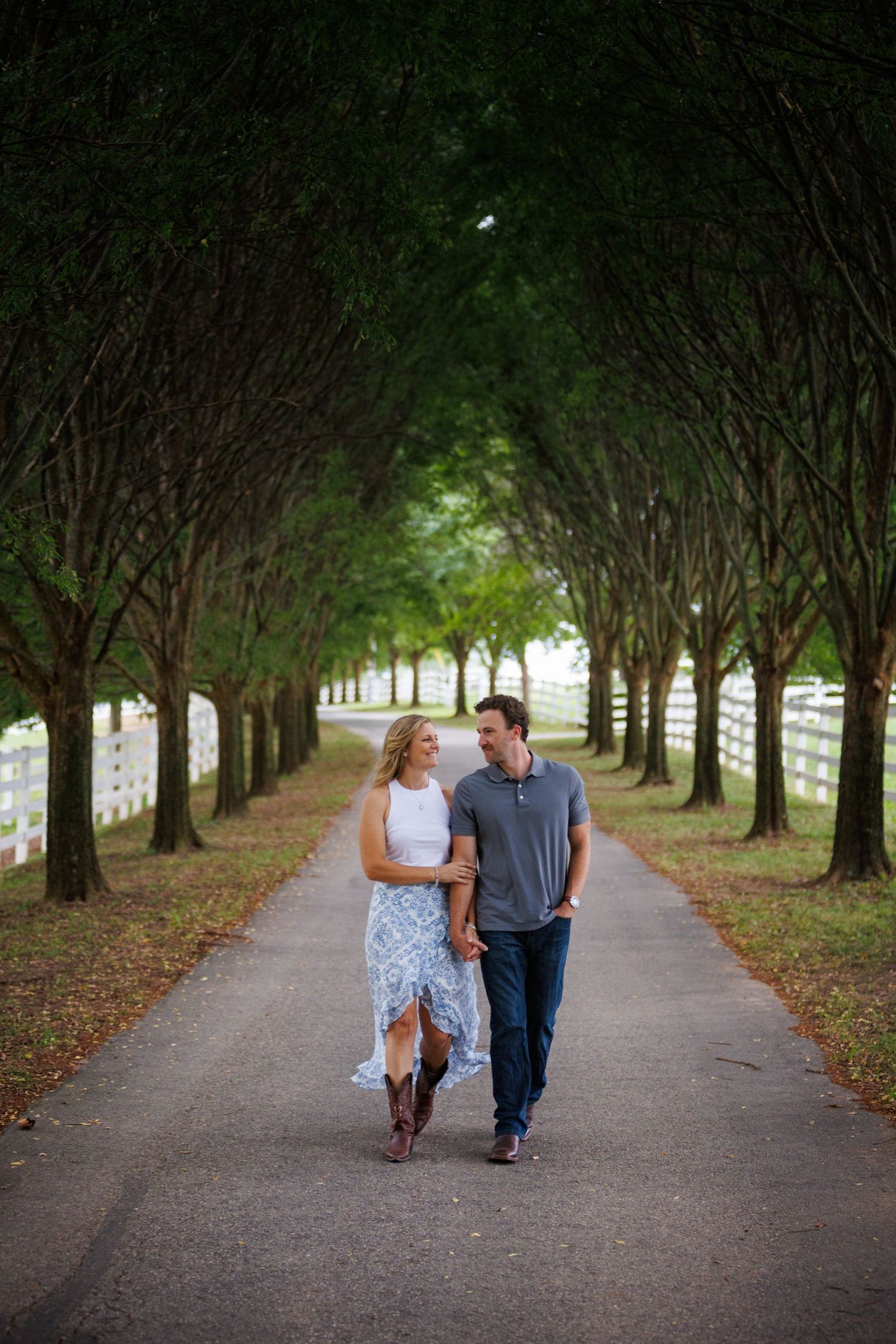 Couple walking hand in hand down a tree-lined path, smiling at each other, woman in white dress and boots.