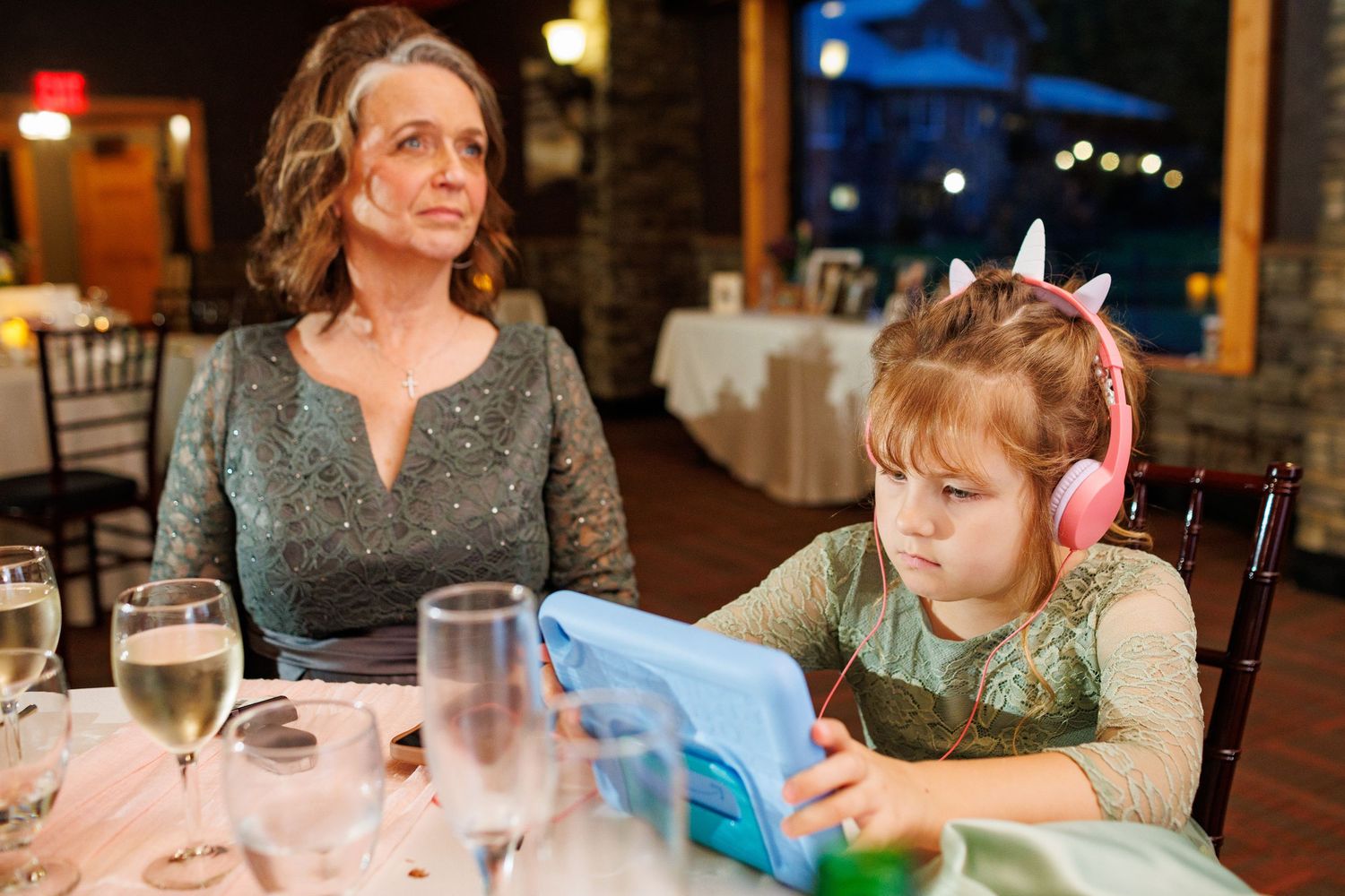 Woman in green dress sits beside young girl wearing pink headphones using tablet at wedding.