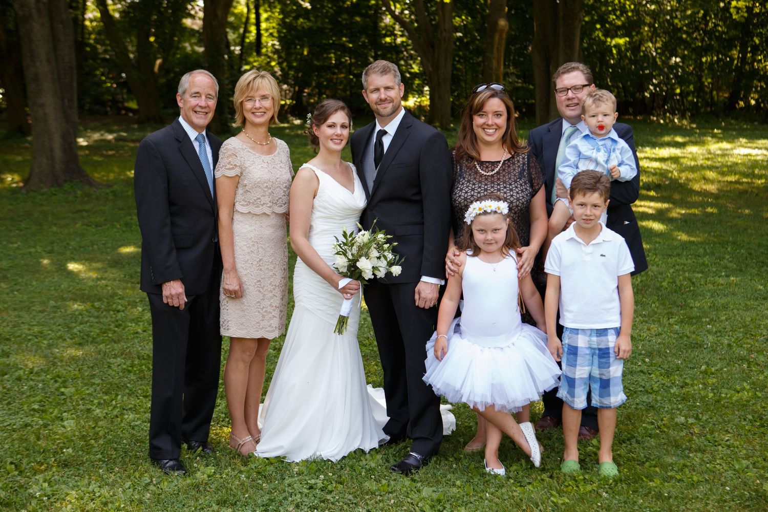 A group poses together outdoors for a formal wedding photo on a sunny day with trees in the background.