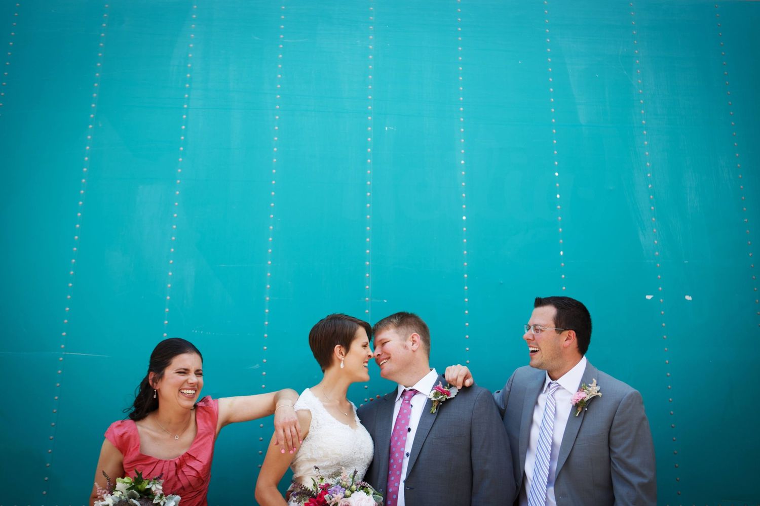 Wedding party posing playfully against vibrant turquoise wall with hanging string lights.