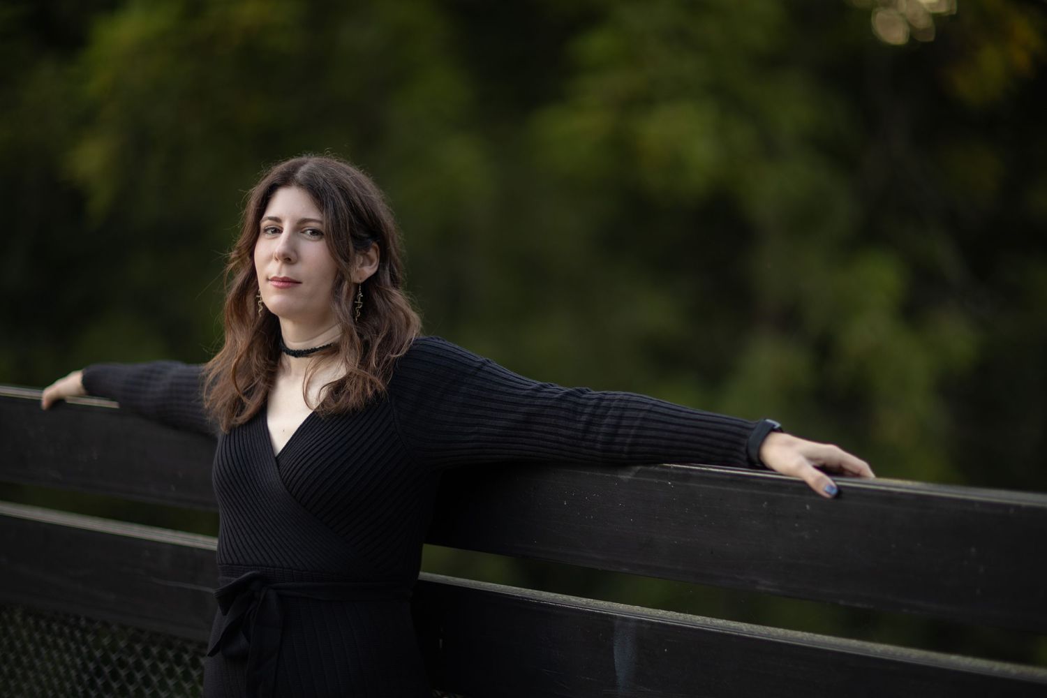 A pensive figure in black attire stands by an industrial railing during an overcast day outdoors.