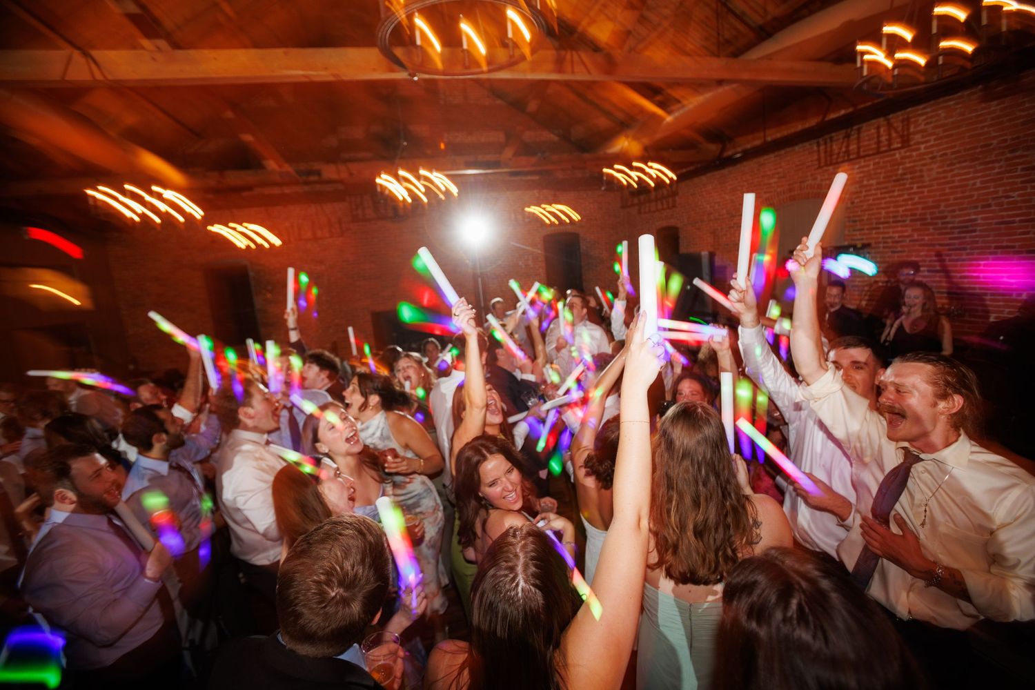 Colorful wedding reception dance floor filled with guests waving glowing LED light sticks at night.