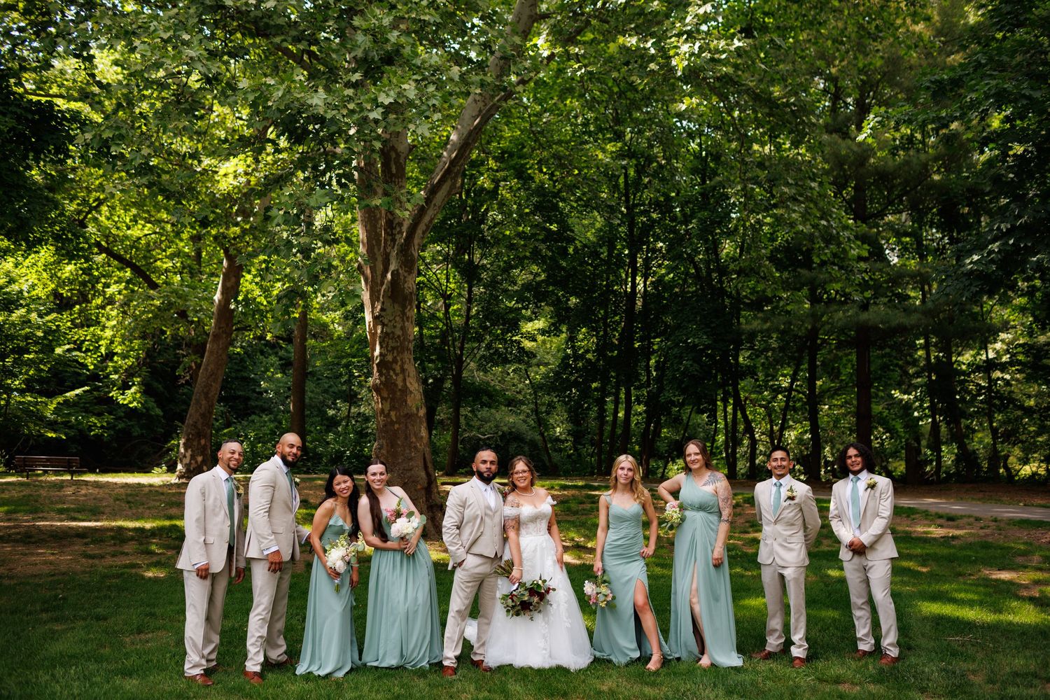 Wedding party poses outdoors in lush green park, bridesmaids in mint gowns and groomsmen in tan suits.