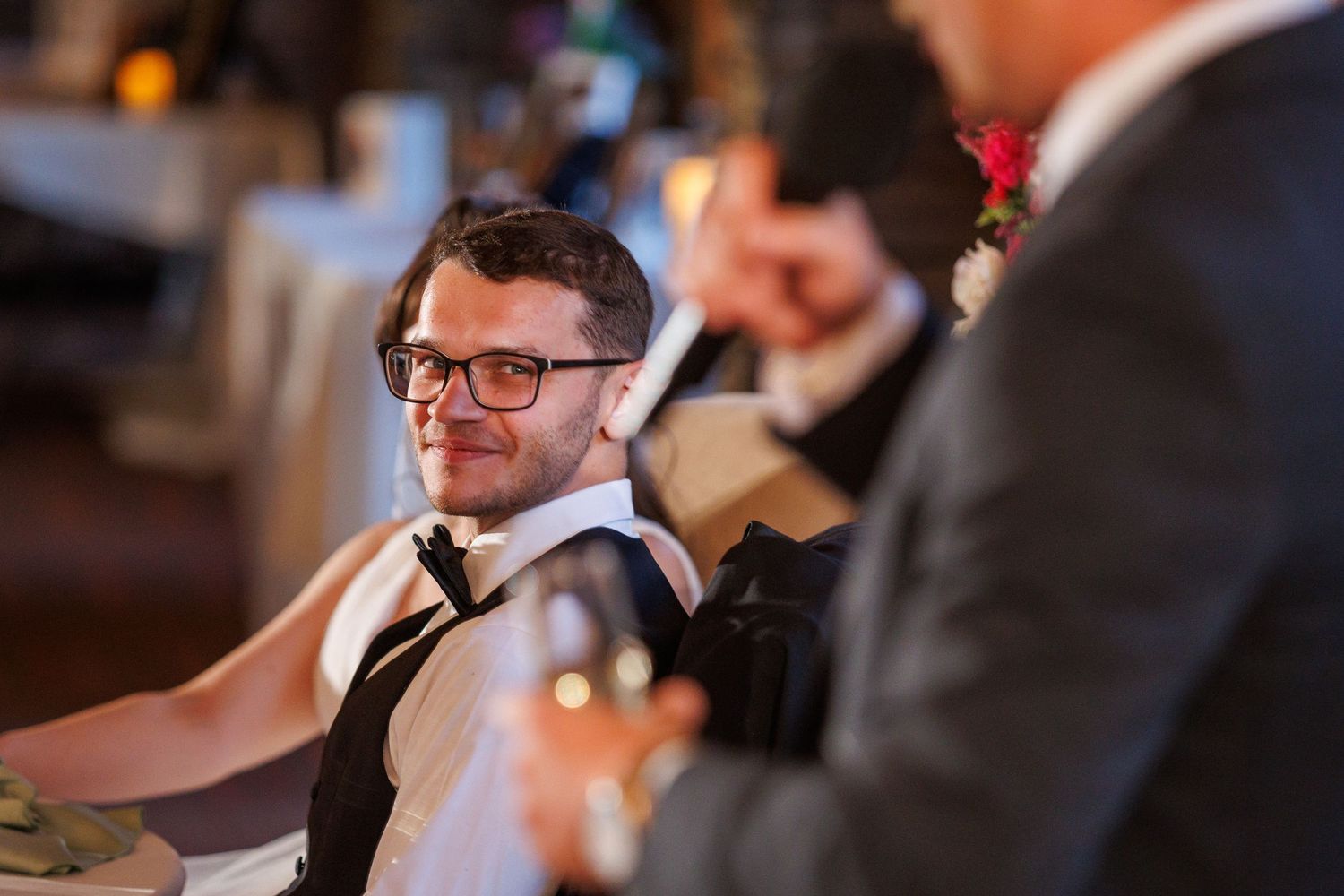 A smiling groom in glasses and bow tie listens to wedding reception speeches.