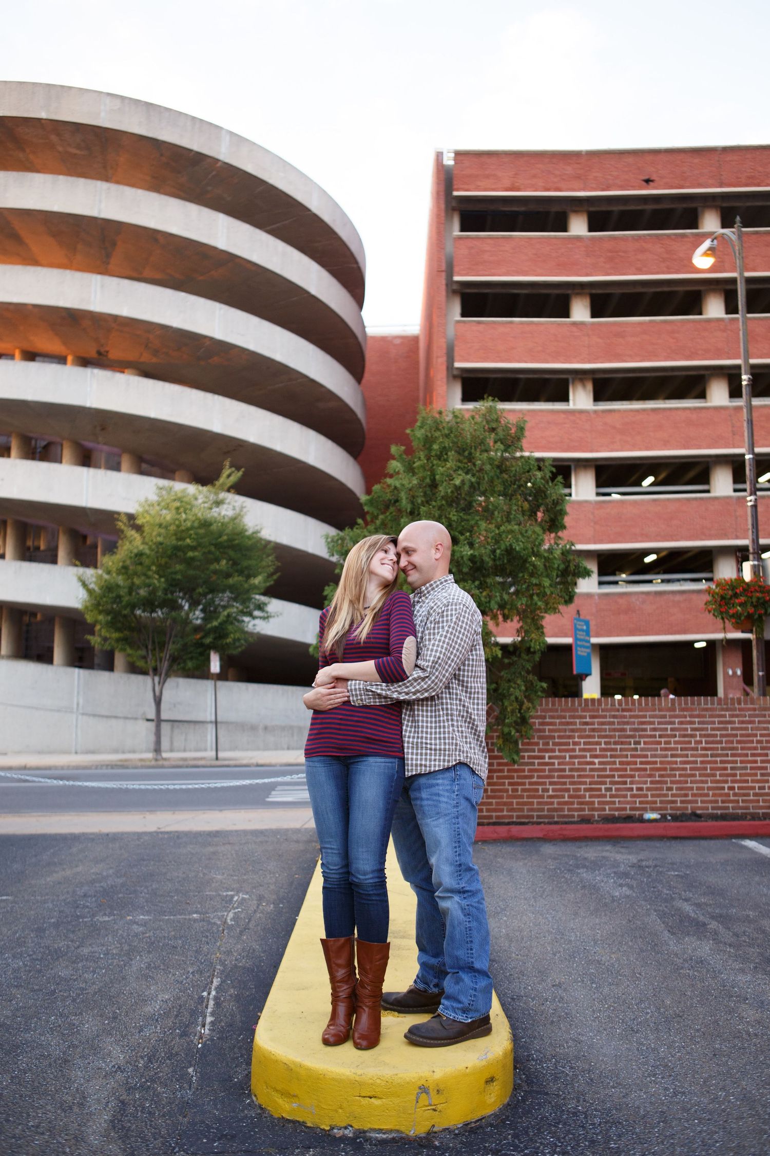 A couple stands together romantically on a yellow painted curb in front of a curved parking garage structure.