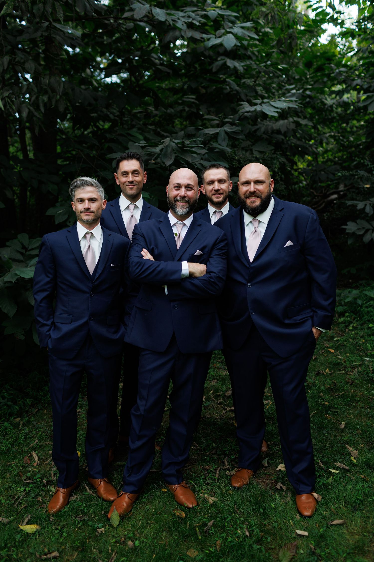 Groomsmen in navy suits and brown shoes pose together in a woodland setting.