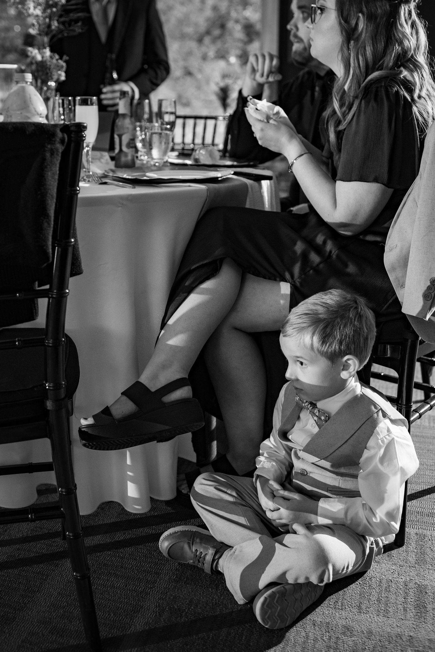 Young boy in suspenders sits on floor eating during wedding reception in black and white photo.
