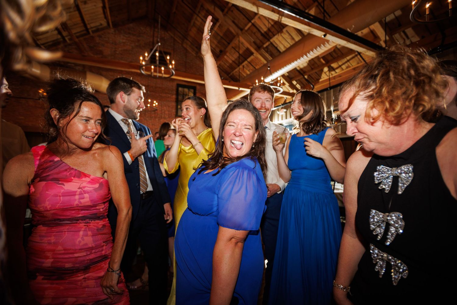 Joyful wedding guests in colorful dresses dance and celebrate at a rustic reception hall.