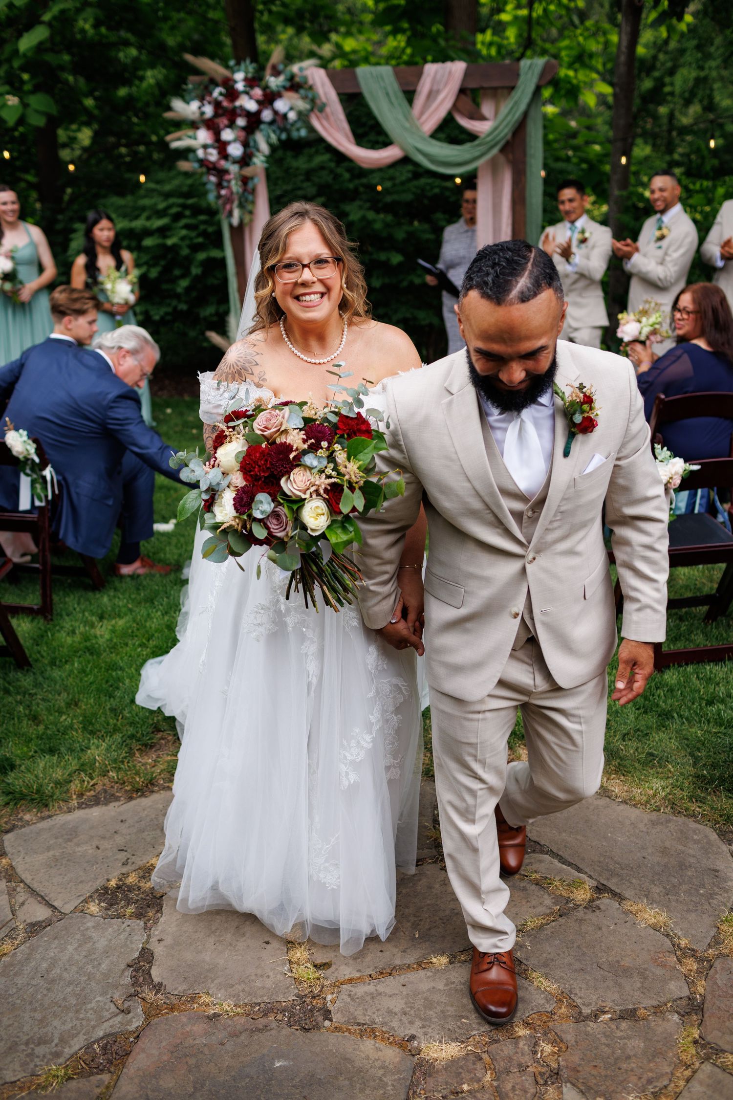 Smiling bride and groom walk down aisle after outdoor wedding ceremony holding bouquet.