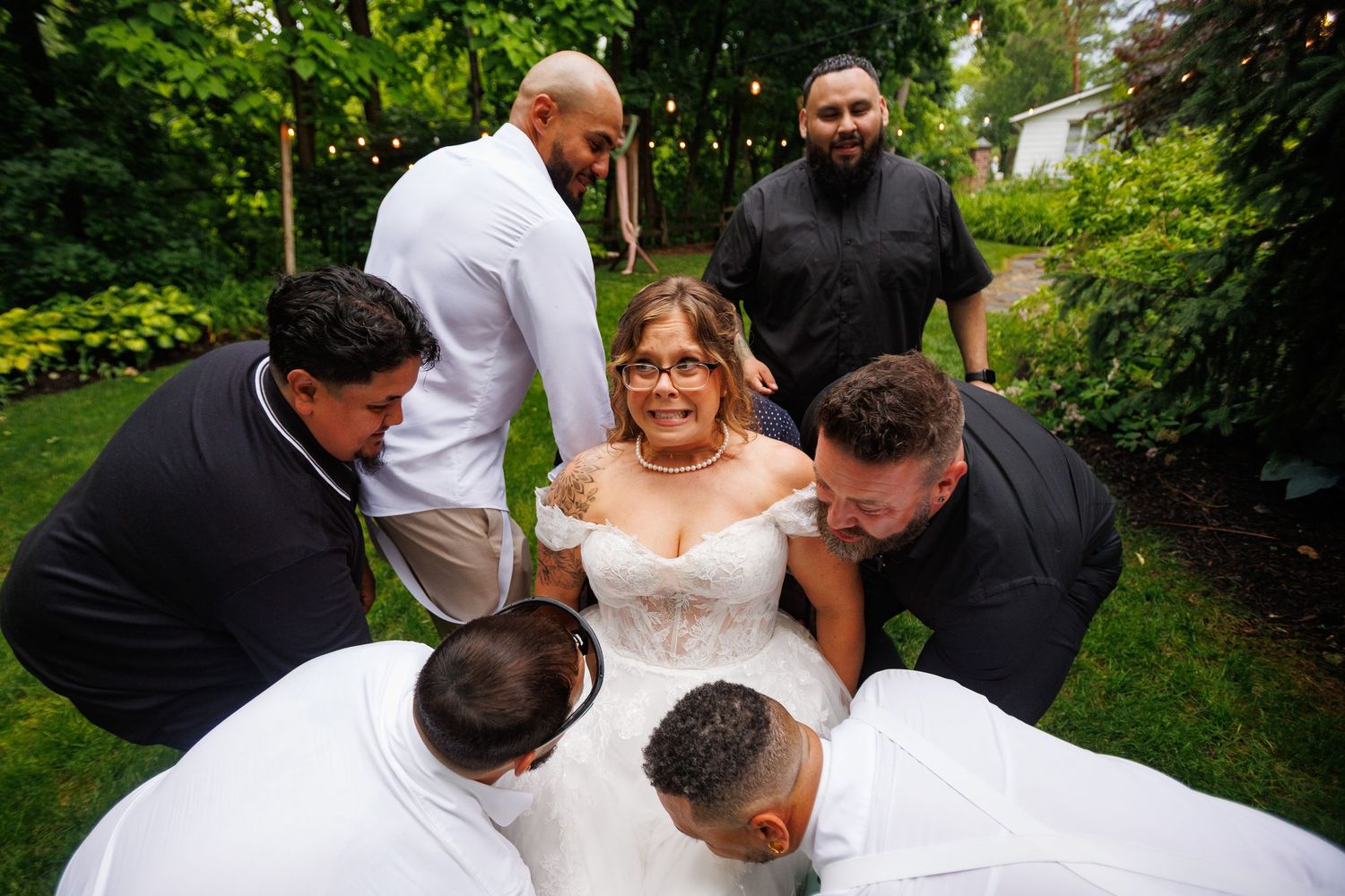 Bride in white gown surrounded by groomsmen in black and white attire during outdoor wedding ceremony.