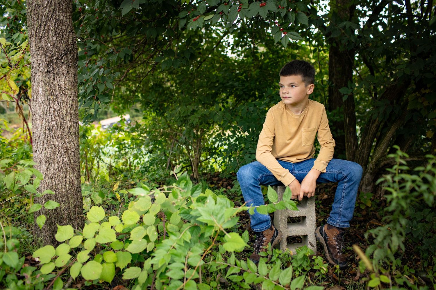 A child in a mustard-colored shirt sits thoughtfully on a wooden chair among green foliage in a garden setting.