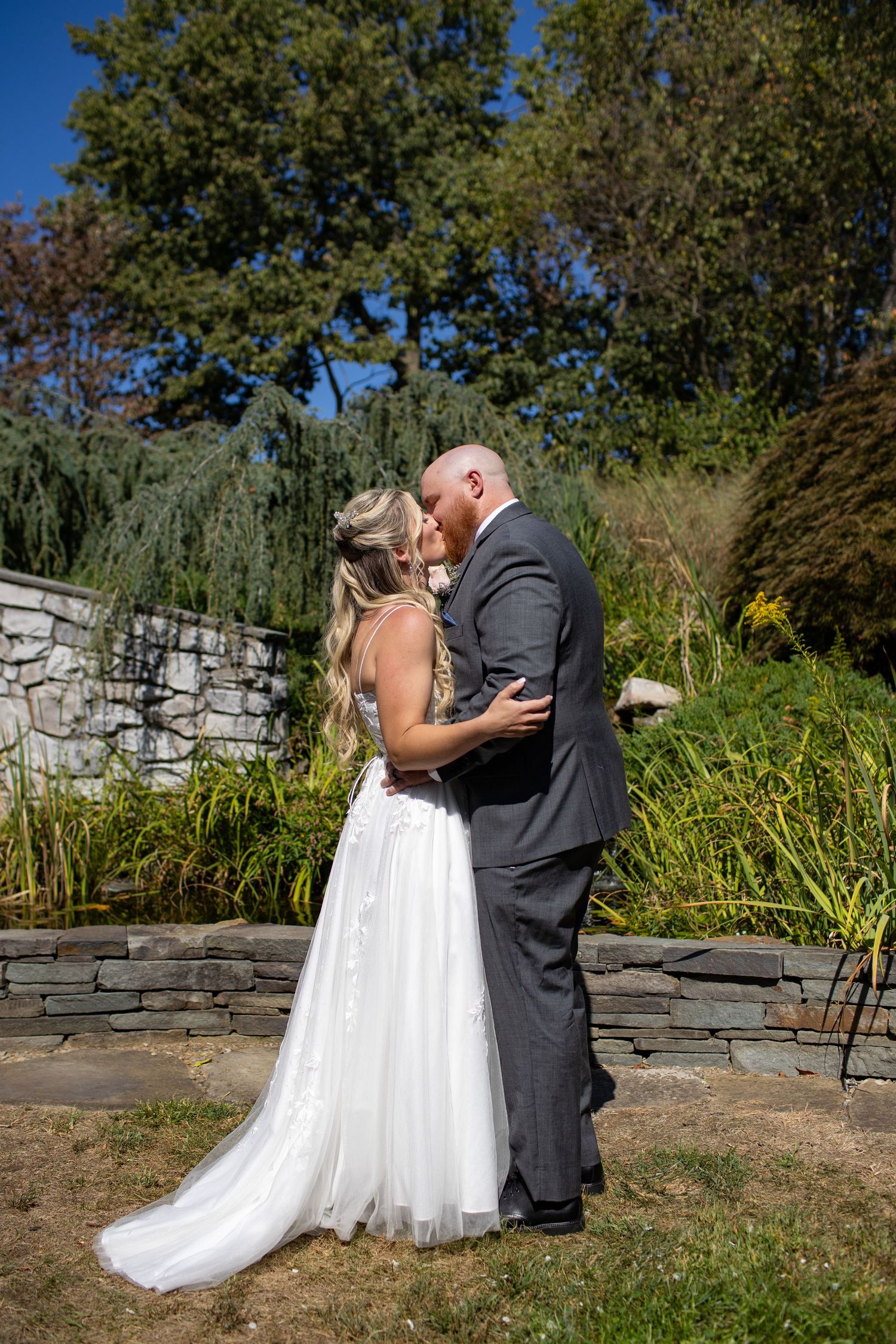 A couple share a romantic moment at a beautiful garden setting with stone walls and lush greenery in the background.