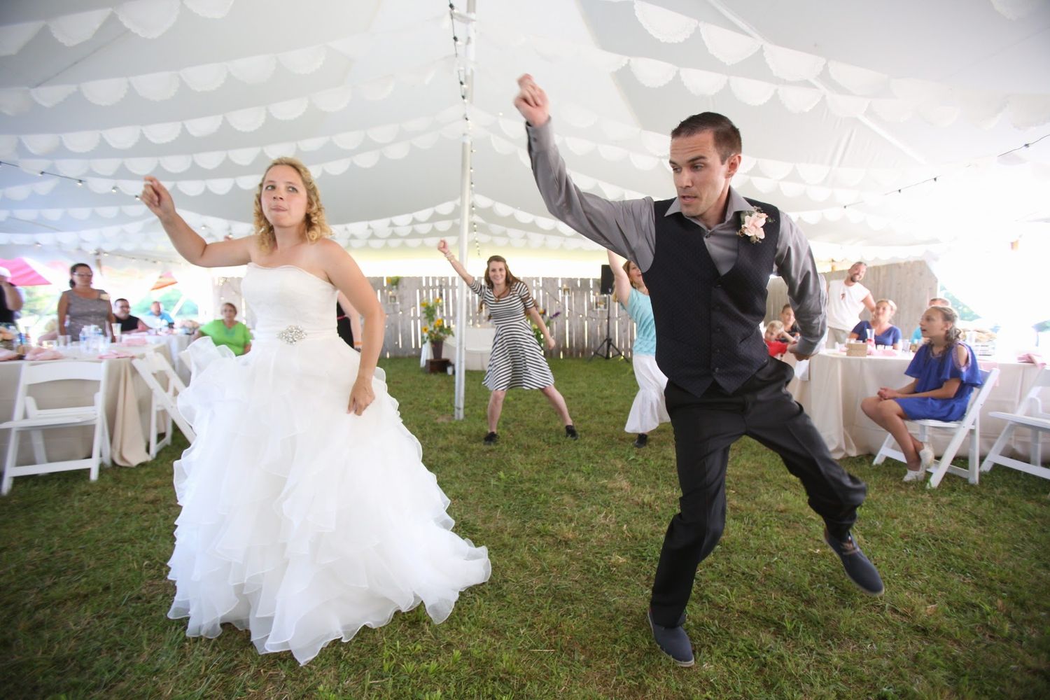 Joyful couple dances energetically at outdoor wedding reception under white tent with guests seated at tables.