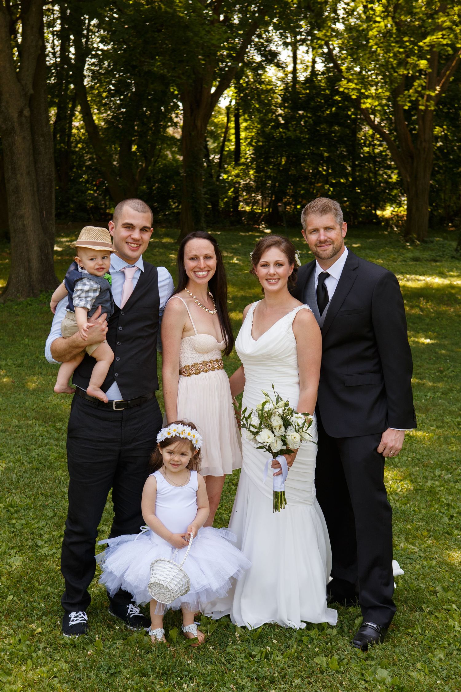 A wedding party poses outdoors in formal attire on a sunny day with trees and greenery in the background.