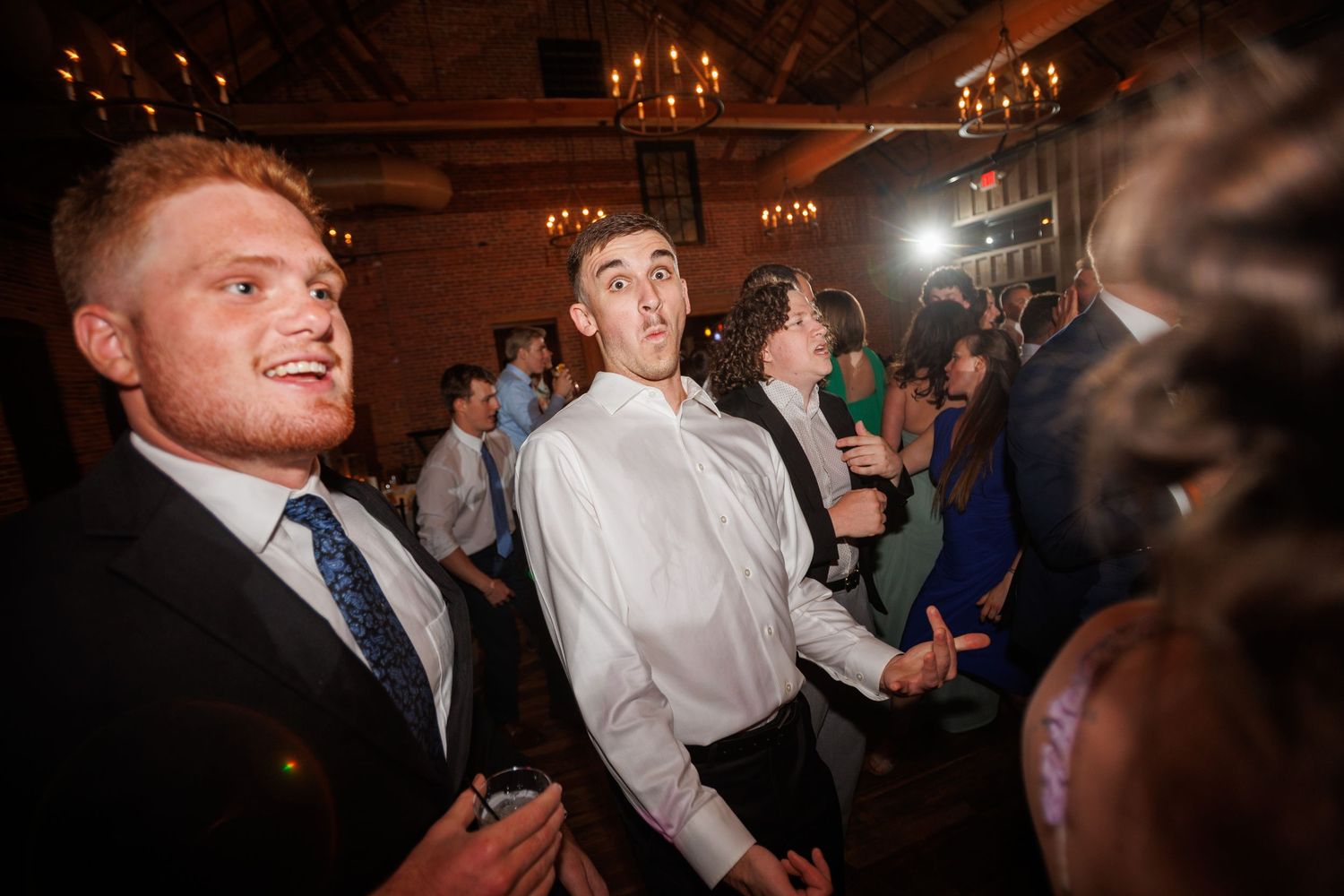 Two young men in suits dance and socialize at a lively wedding reception in a warmly lit venue.
