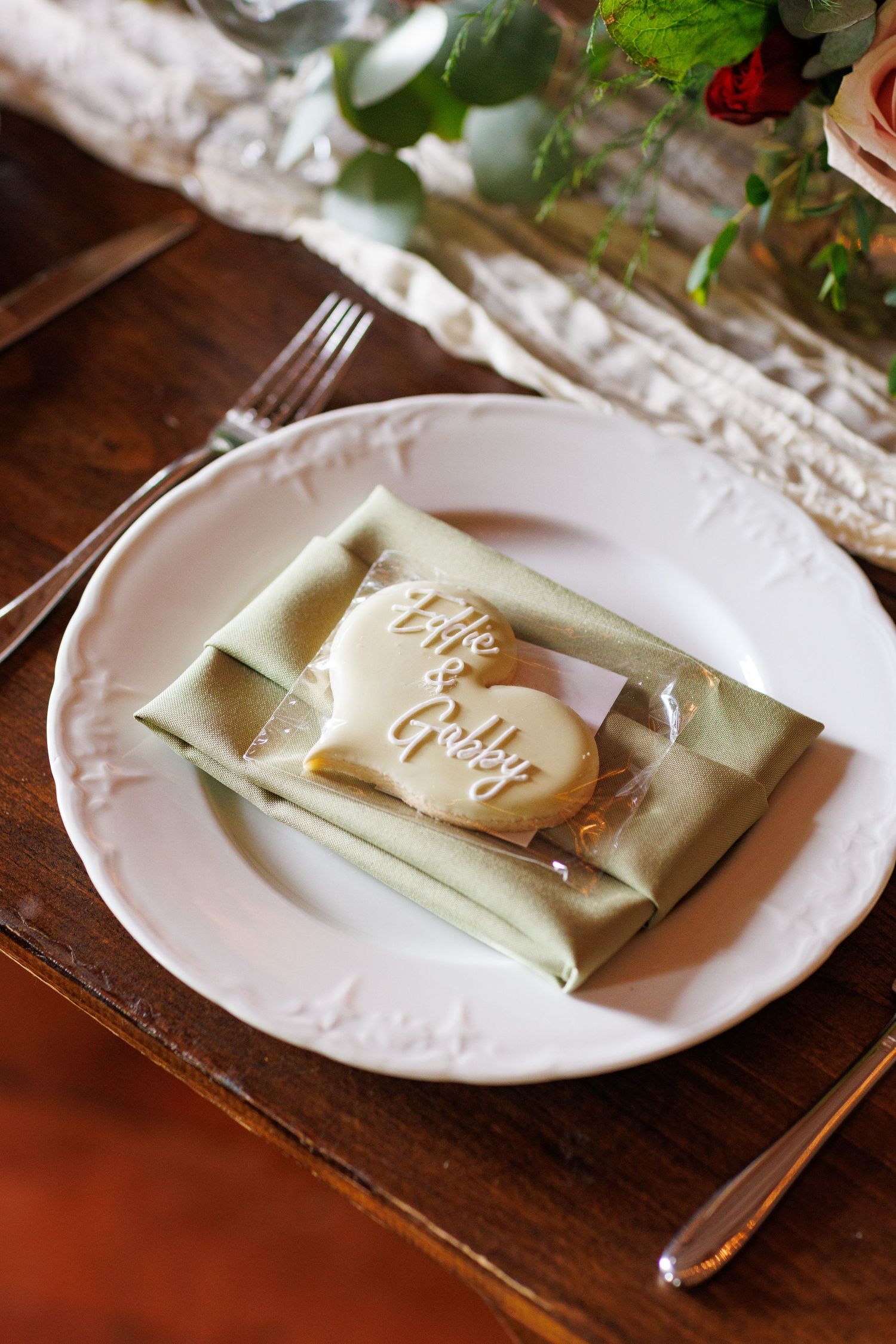Elegant wedding place setting with a heart-shaped cookie inscribed with couple names on a sage green napkin.