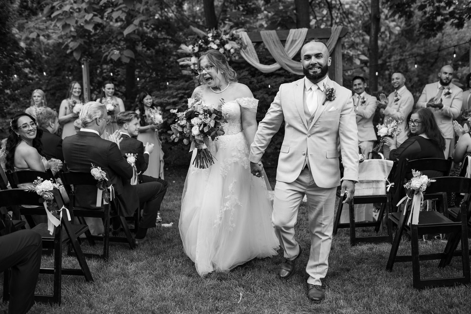 Newlyweds joyfully walk back down the aisle after outdoor wedding ceremony in black and white photo.